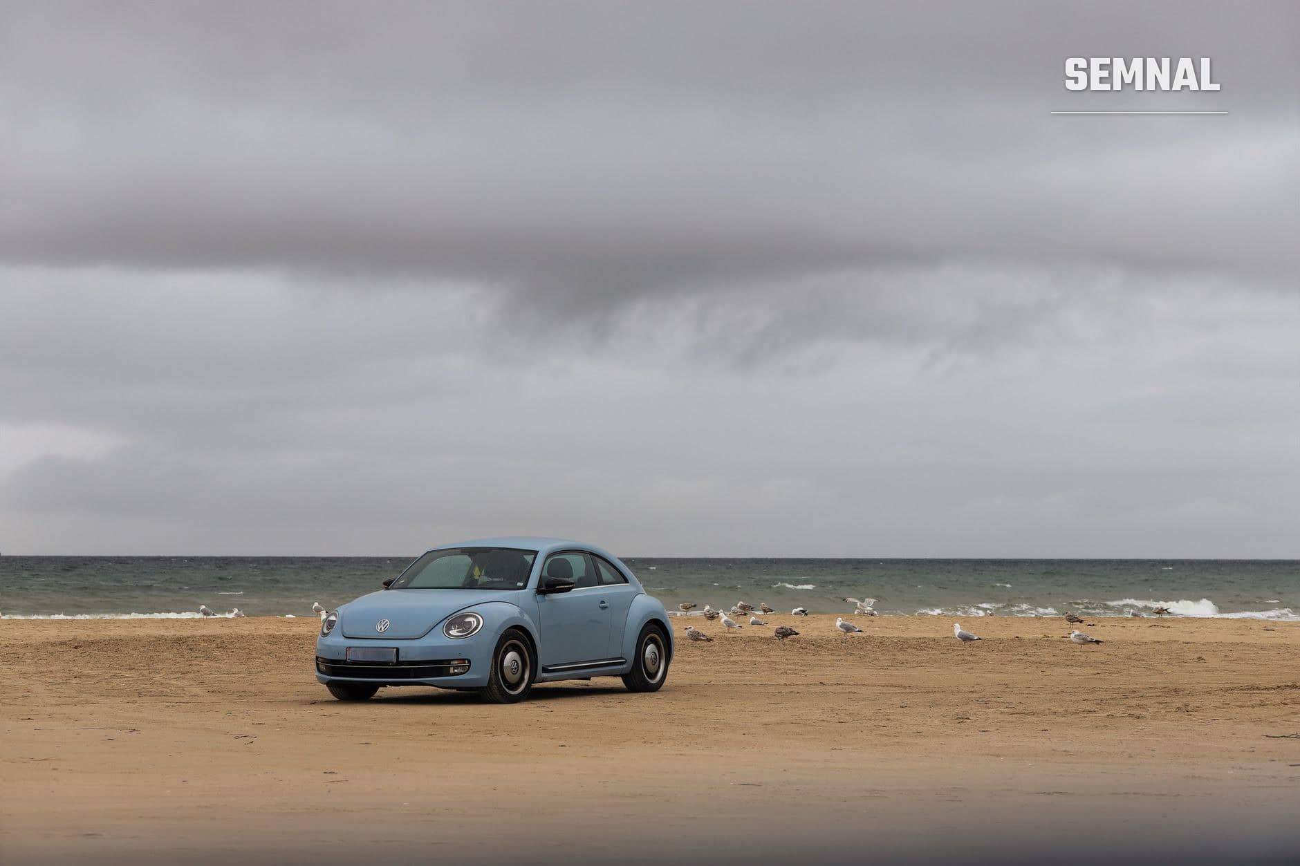 Volkswagen Beetle albastru parcat pe nisipul gol al unei plaje, cu marea agitată și cerul noros în spate, ilustrând incidentul de amendaracomandanți. (Foto: Christina & Peter / Pexels)
