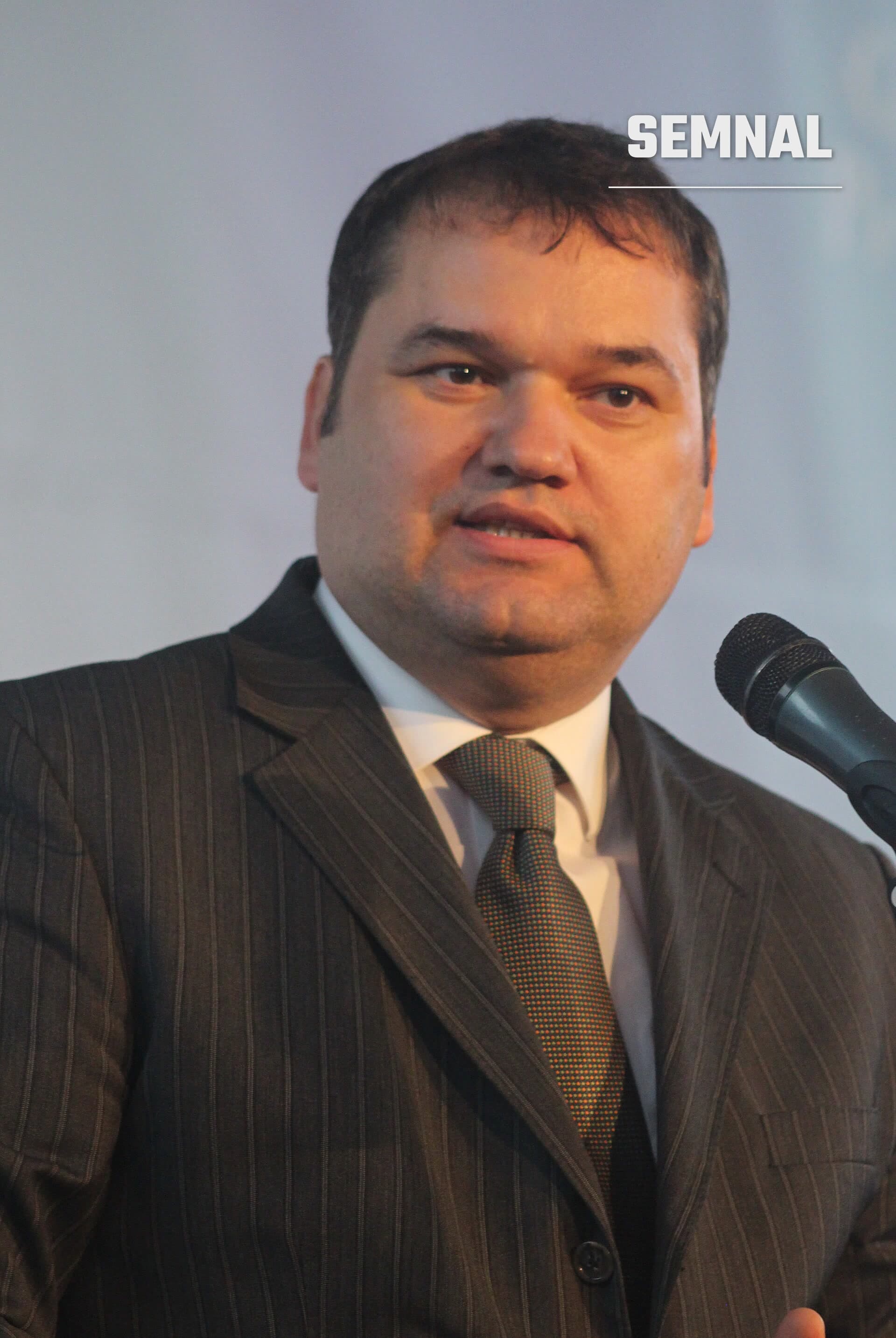 Cseke Attila vorbind la microfon, într-un costum cu dungi, cămașă albă și cravată cu model, pe un fundal gri-albăstrui. (Foto: László Horváth)
