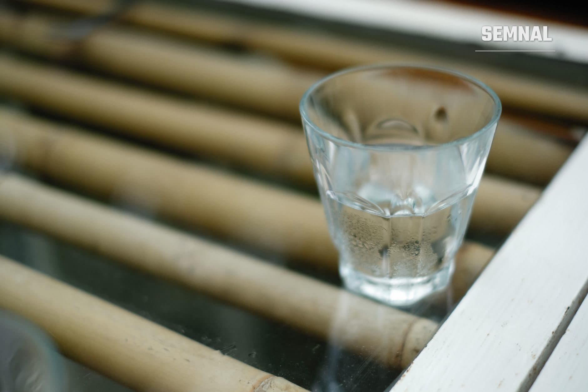 Un pahar transparent cu apă pe o suprafață de bambus, cu fundal estompat și un cadru alb de lemn în colțul din dreapta. (Foto: Alexey Demidov / Pexels)