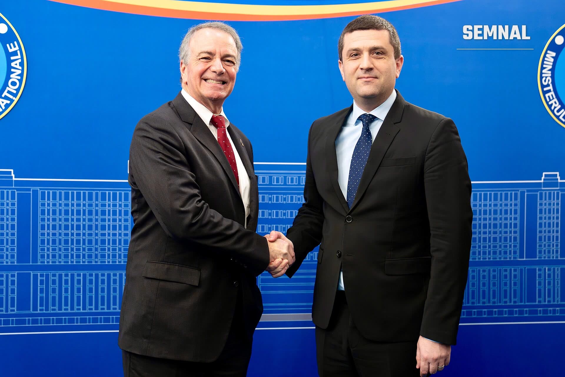 Radu Miruță strânge mâna unui alt bărbat în costum închis, în fața unui fundal albastru cu inscripții oficiale. (Foto: Photos by U.S. Embassy Romania)