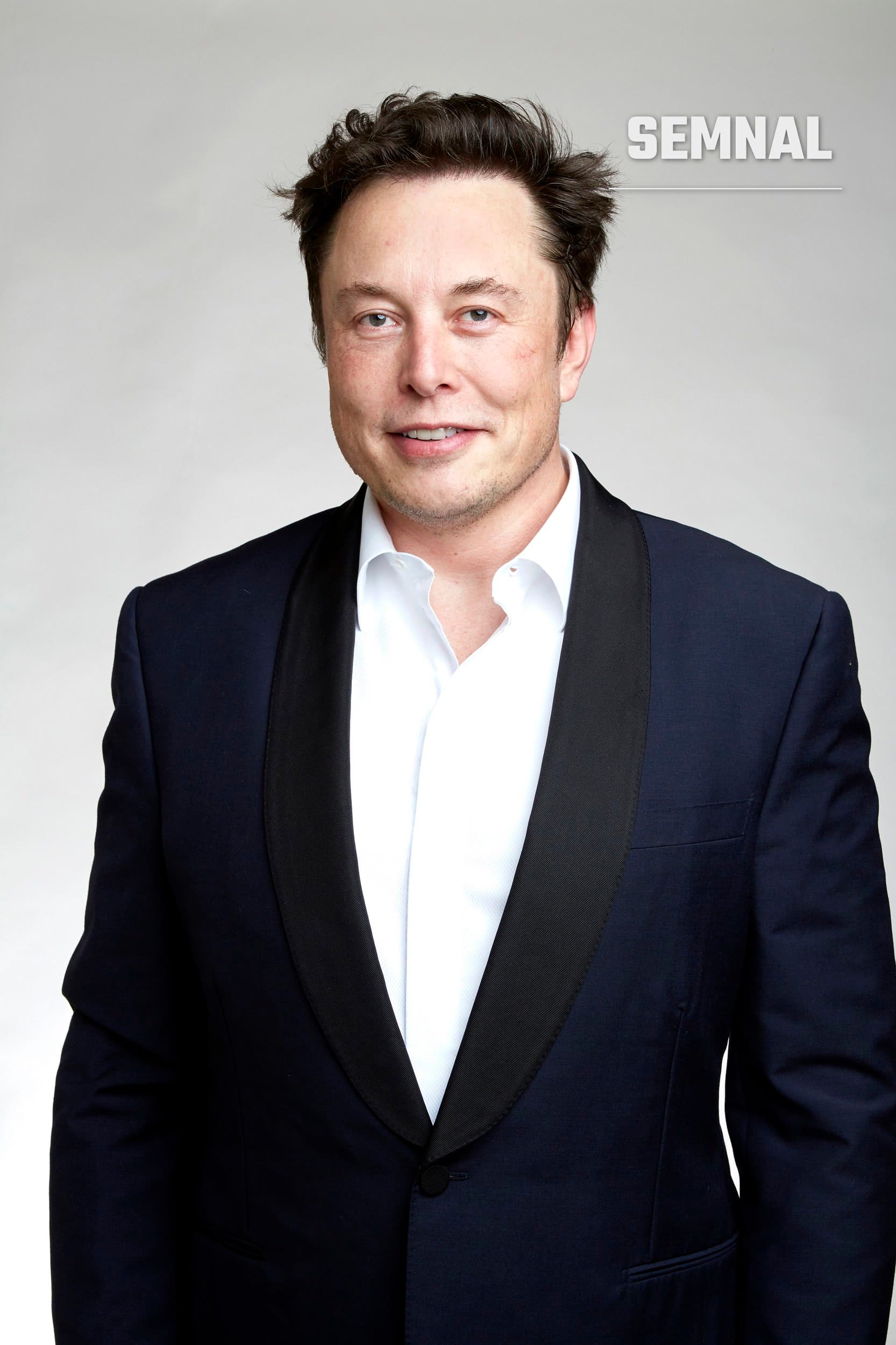 Elon Musk, într-un portret de studio pe fundal gri, zâmbind discret, în costum bleumarin cu revere negre și cămașă albă. (Foto: Duncan.Hull)