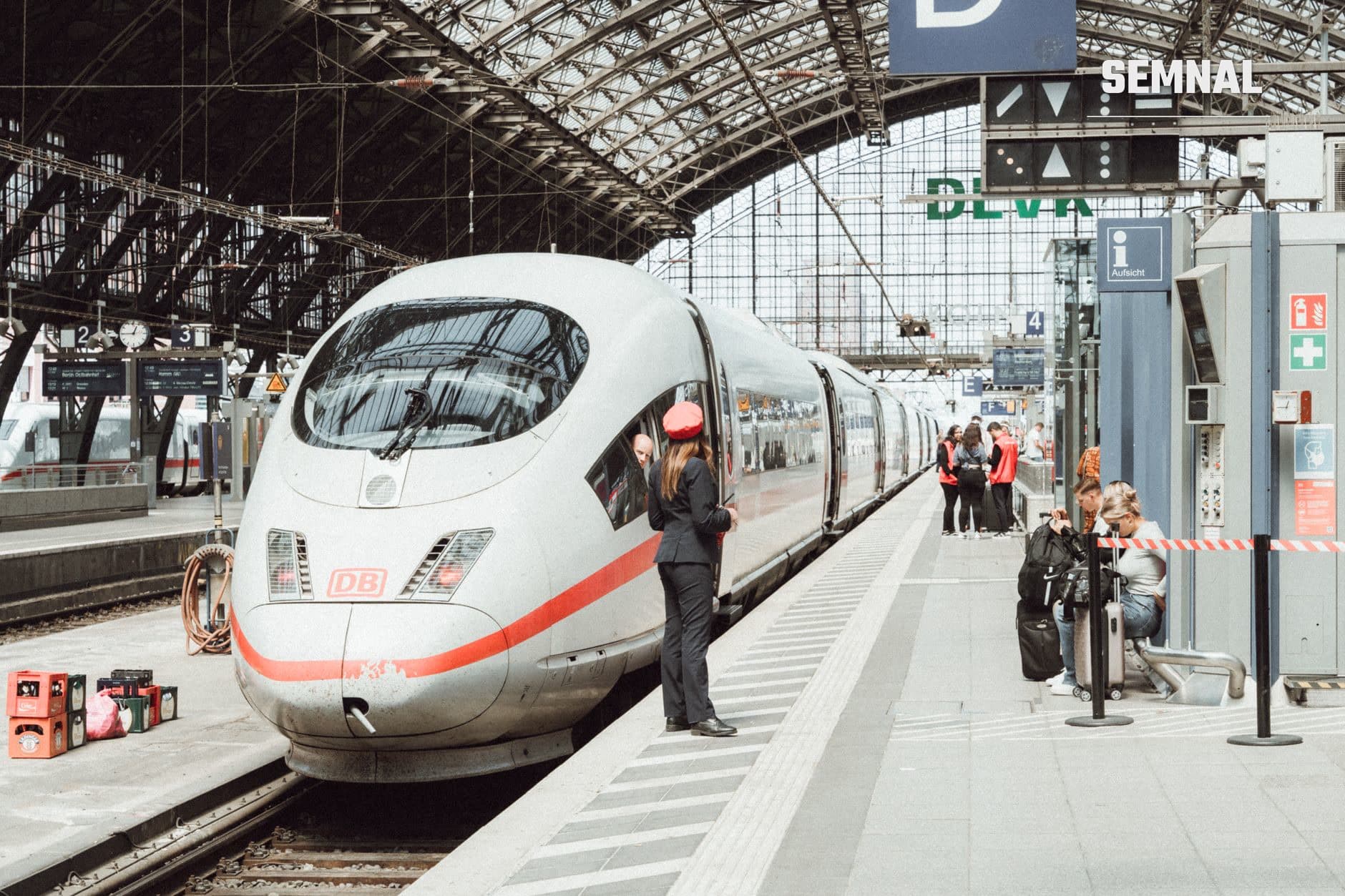 Trenul ICE alb cu dungă roșie, oprit la peron într-o gară cu acoperiș metalic și panouri de plecări. (Foto: Masood Aslami / Pexels)