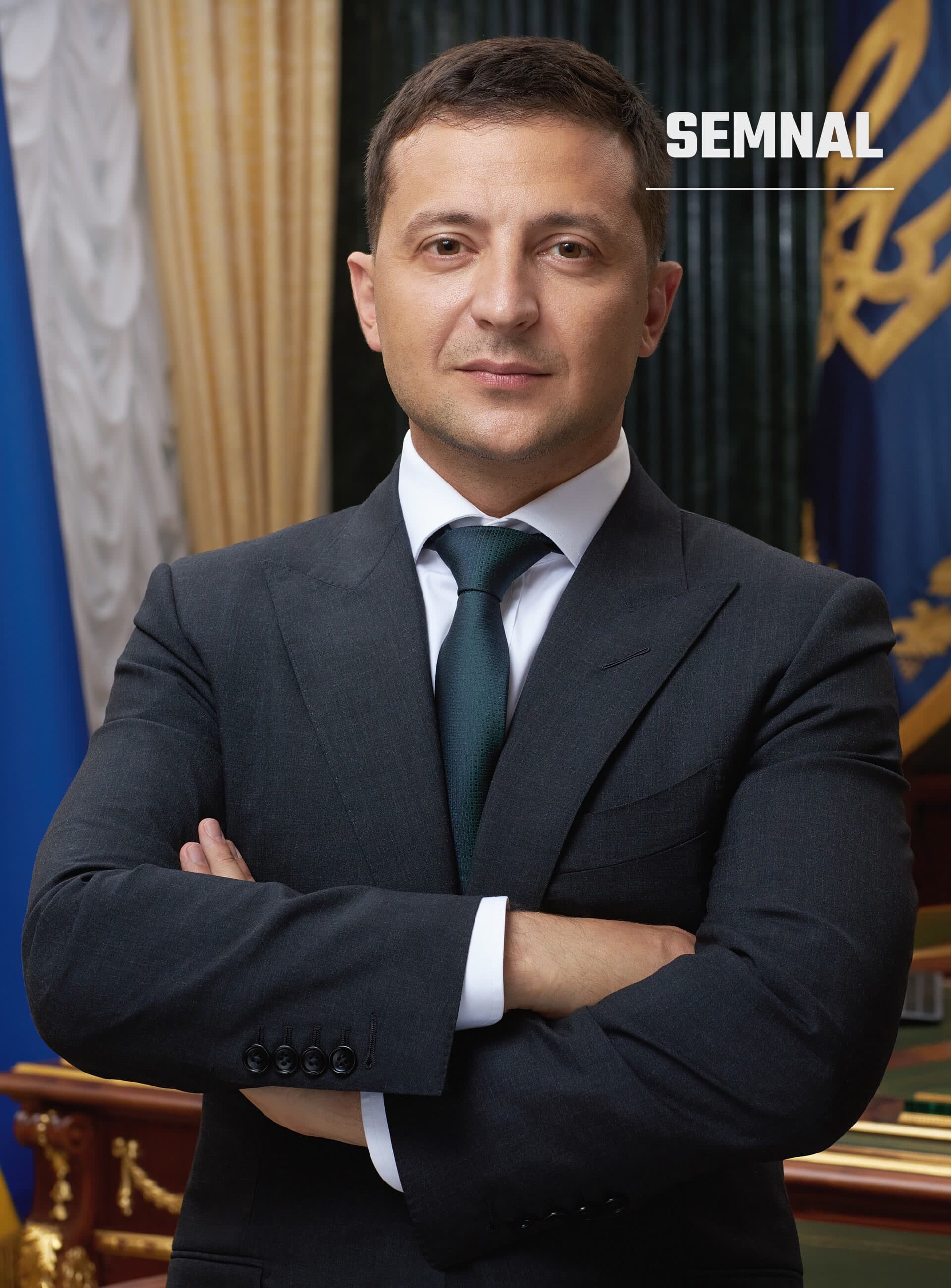 Volodimir Zelenski, în costum gri inchis și cravată verde, cu brațele încrucișate, în fața unor steaguri și draperii aurii. (Foto: http://www.president.gov.ua/)