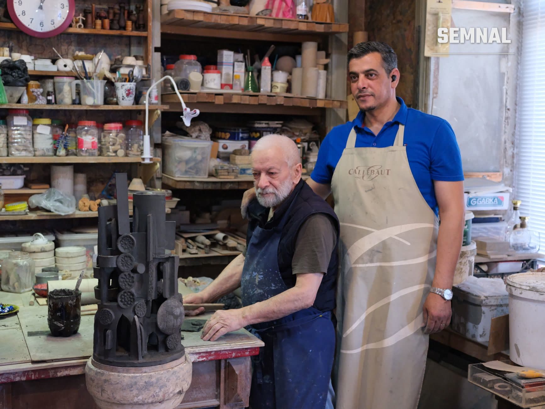 Un meșter în vârstă modelează o sculptură din lut într-un atelier de ceramică, alături de un bărbat tânăr în șorț bej. (Foto: Tahir Xəlfəquliyev / Pexels)