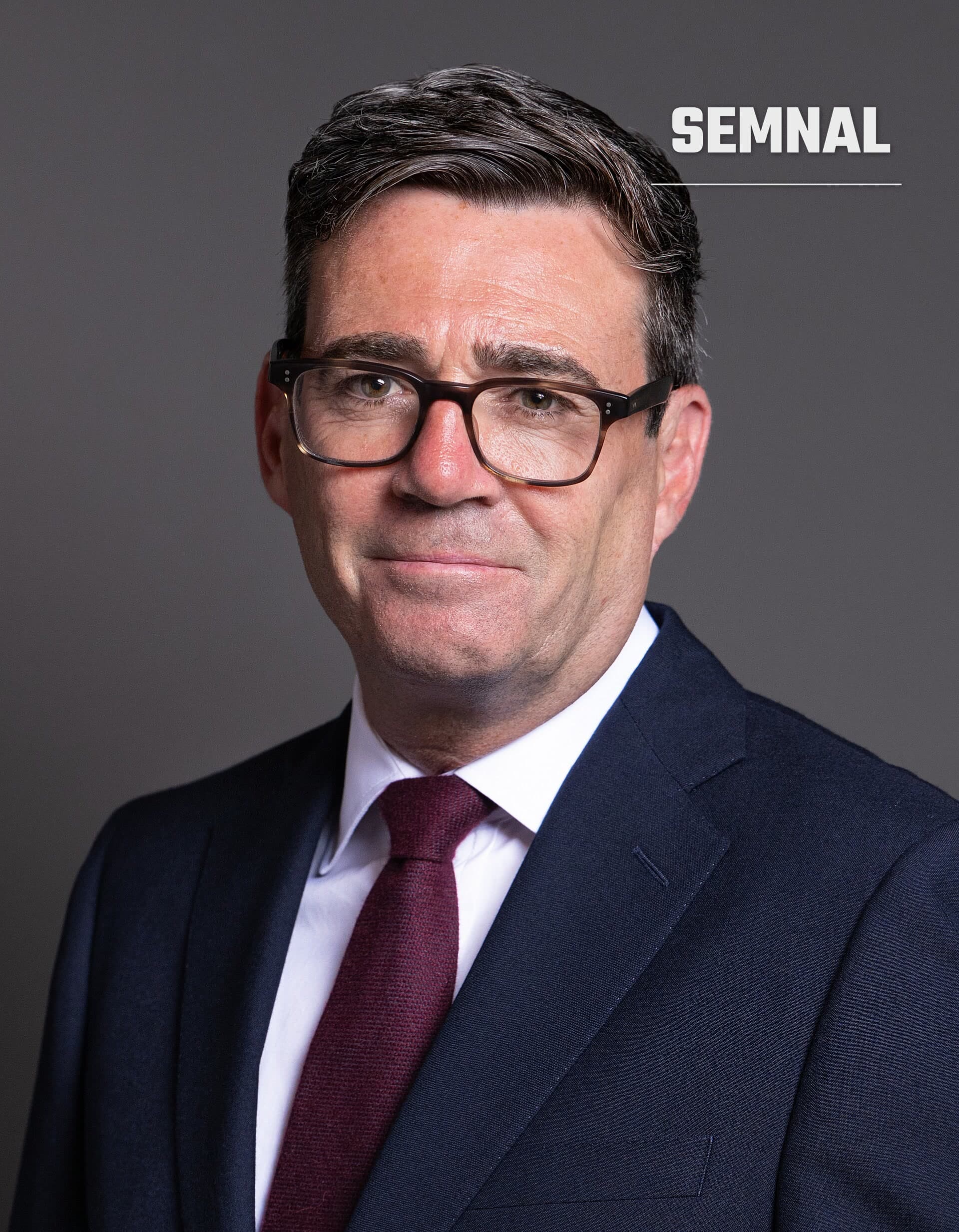 Andy Burnham, în portret oficial, cu ochelari cu rama maro, cămașă albă și cravată bordo, pe fundal gri. (Foto: House of Commons)