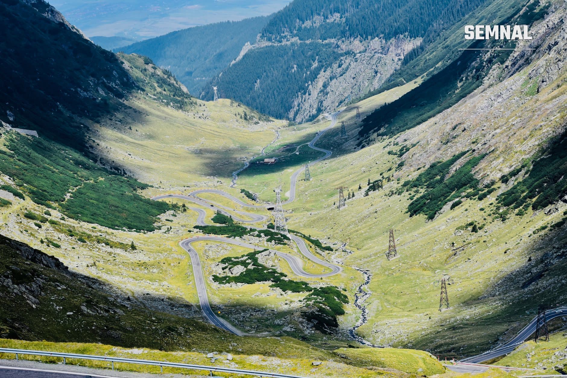 Șosea de munte cu serpentine întortocheate printr-o vale alpină verde, cu stânci, pâraie și stâlpi de înaltă tensiune. (Foto: Chiper Catalin-Marian / Pexels)