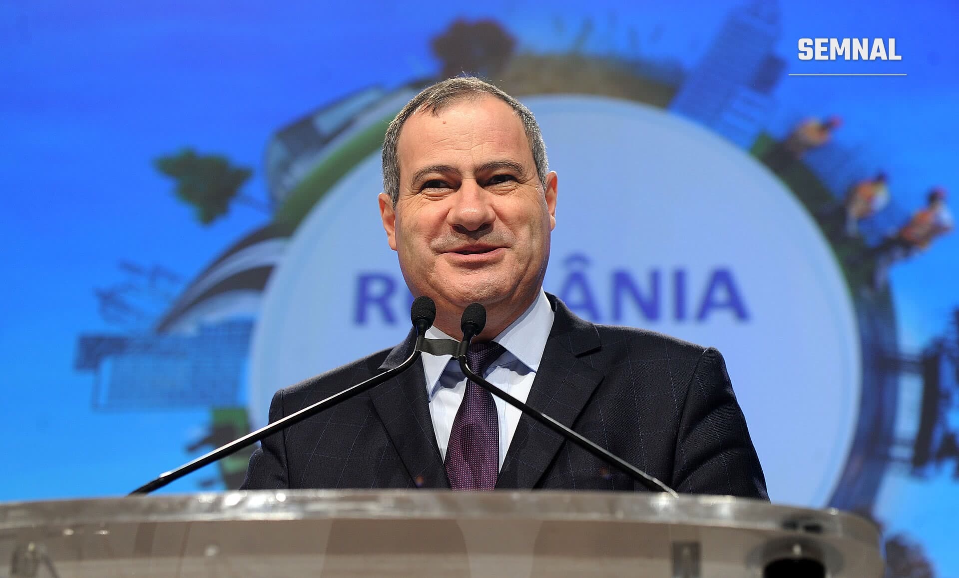Marian Neacșu vorbește la un pupitru cu microfoane, în fața unui ecran albastru cu textul România, zâmbind, în costum închis și cravată mov. (Foto: Partidul Social Democrat)