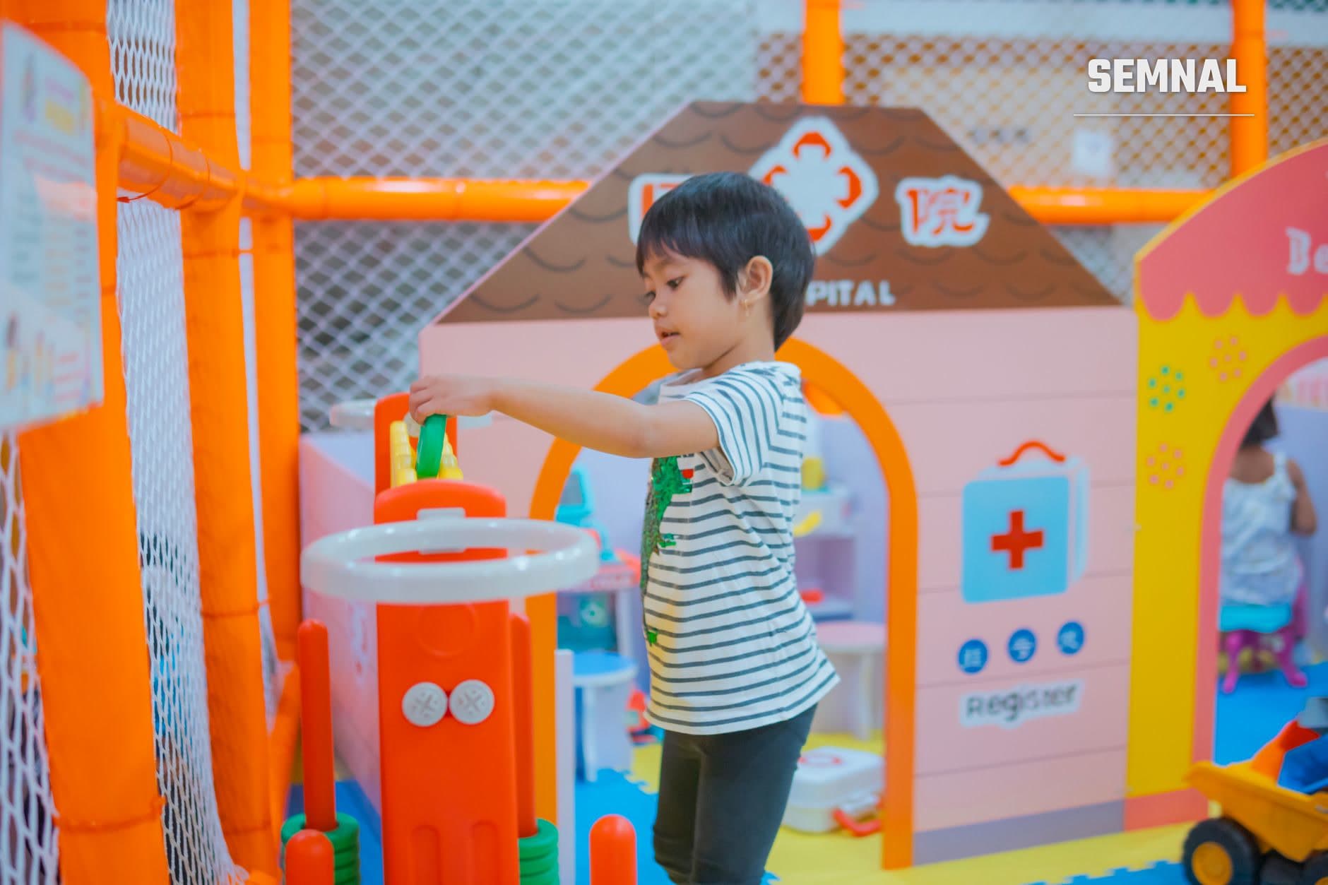 Un copil se joacă într-un spațiu de joacă interior, lângă o căsuță roz tip spital, cu cruce medicală și jucării colorate. (Foto: Nothing Ahead / Pexels)