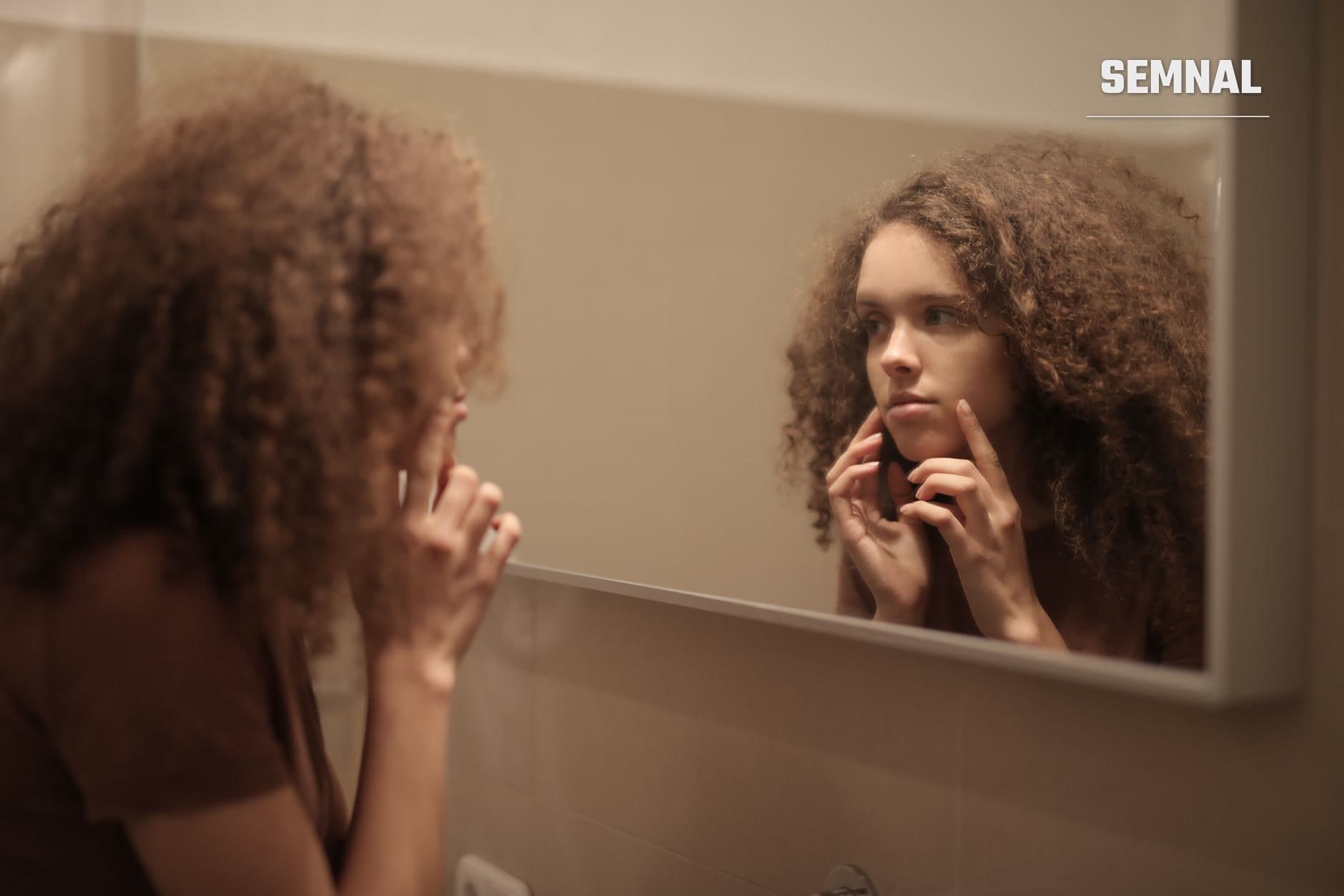 Femeie cu păr creț, castaniu, care se privește în oglinda de baie și își atinge obrazul cu degetele, examinându-și pielea feței. (Foto: Andrea Piacquadio / Pexels)