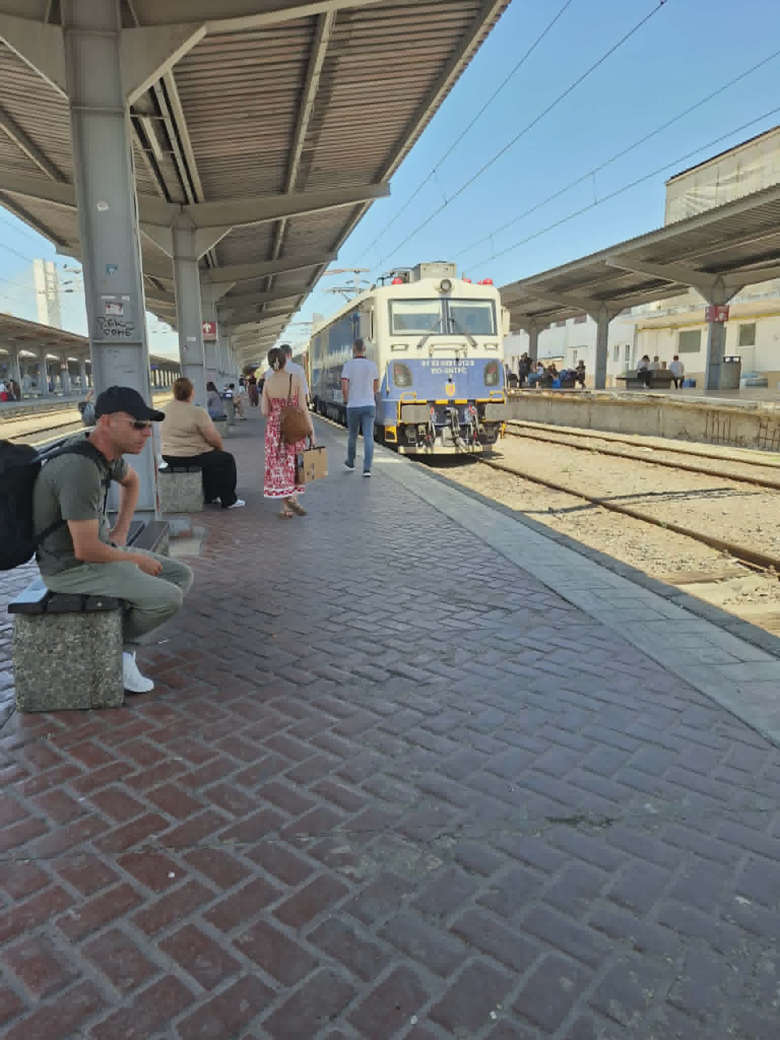 Călători așteaptă pe peronul unei gări, cu un tren oprit la linie