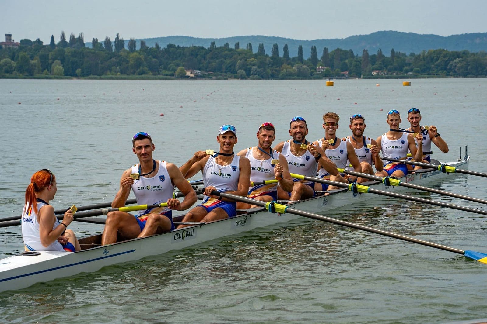 Echipajul masculin de opt plus unu al României pozează cu medaliile de aur în barcă, pe lac, la Campionatele Europene de canotaj de la Varese