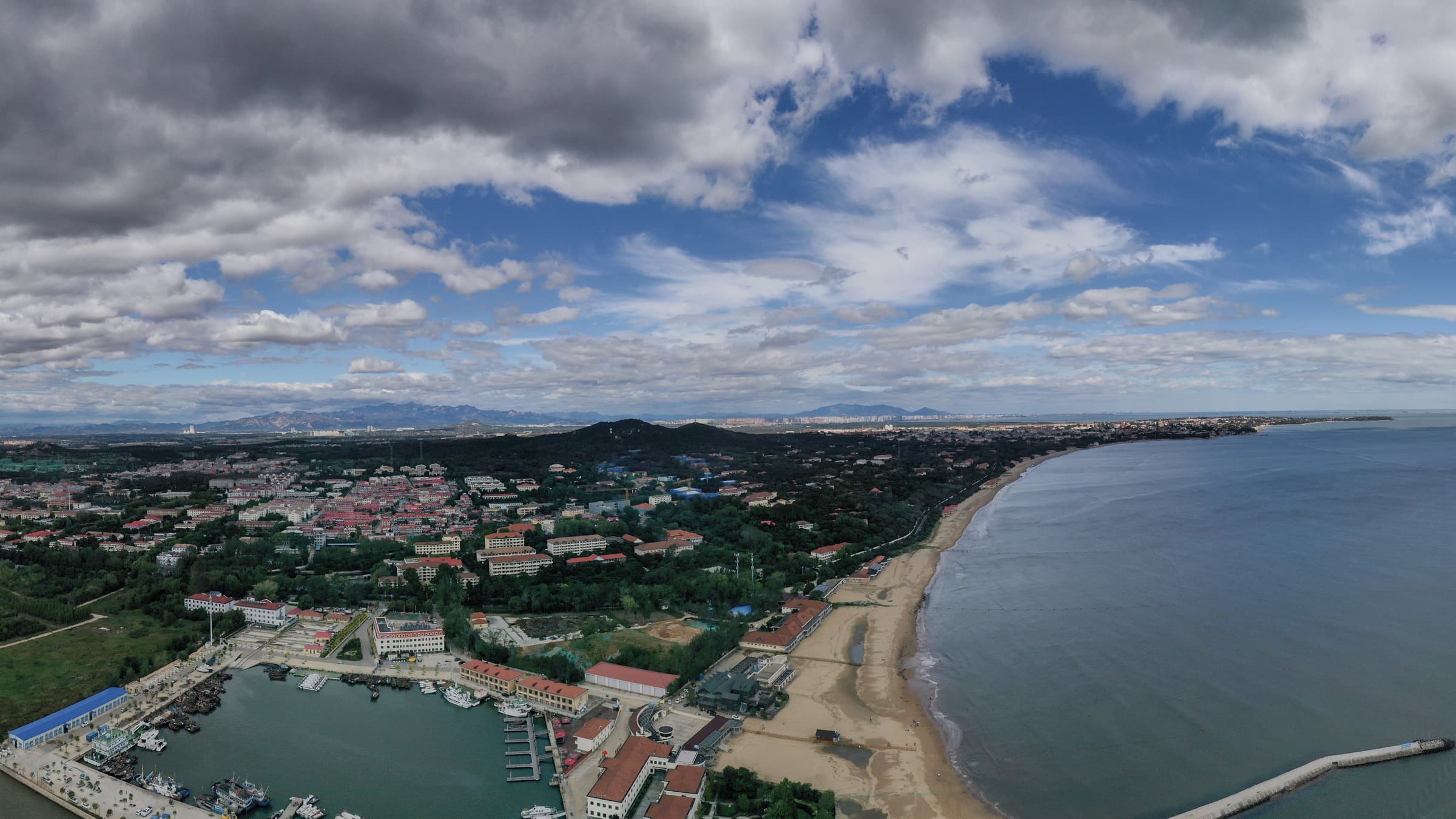 Vedere aeriană asupra stațiunii Beidaihe, cu plaja întinsă de-a lungul golfului Bohai, portul de ambarcațiuni și cartierele cu vile ascunse între copaci