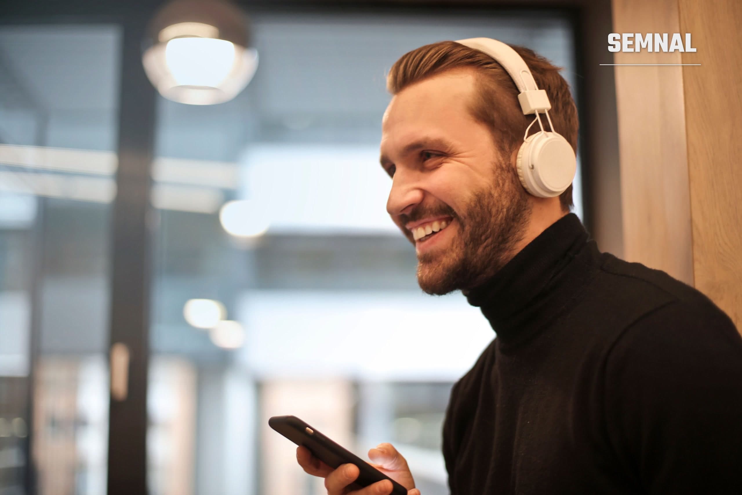 Bărbat cu barbă zâmbind, purtând căști albe și ținând un smartphone, ascultând muzică în interior