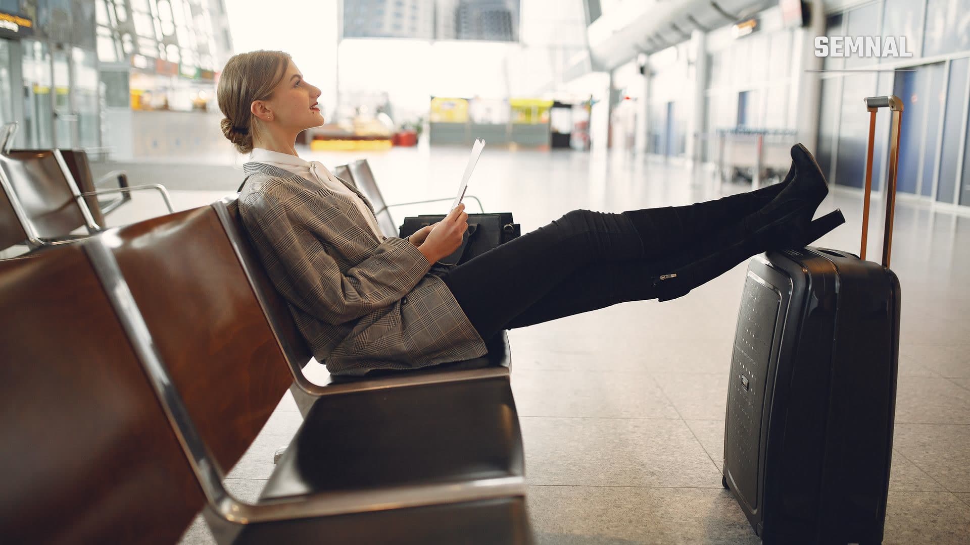 Călătoare relaxată în zona de plecări a unui aeroport, cu picioarele sprijinite pe bagajul de mână, citind un document înainte de îmbarcare