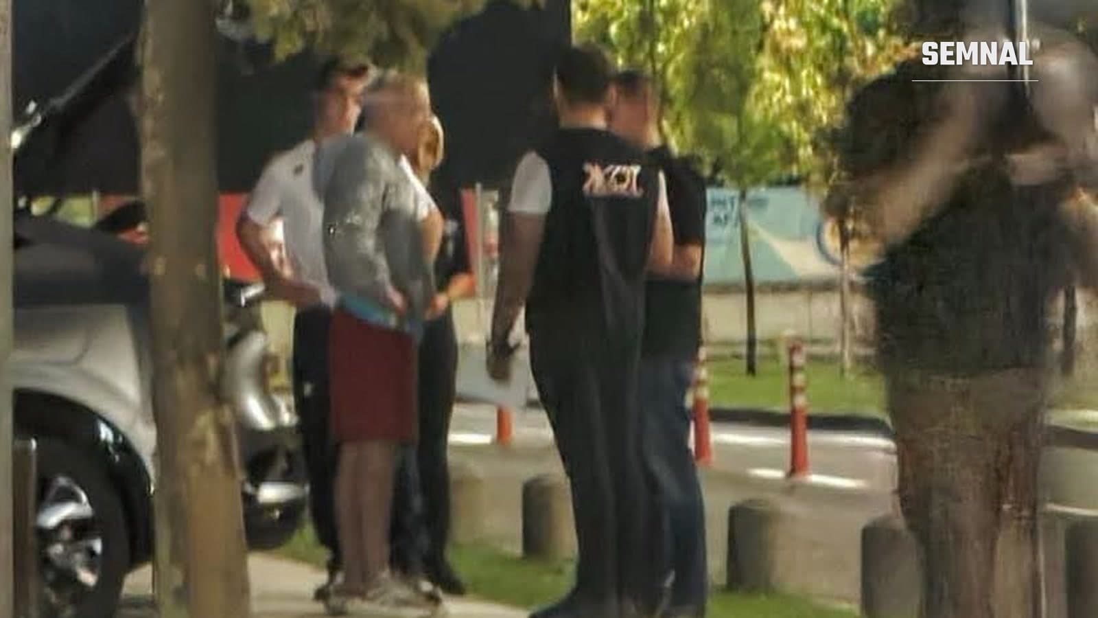 Grup de persoane oprite pe trotuar lângă o mașină, printre ele un bărbat în vestă neagră cu inscripție și un bărbat în cămașă de uniformă