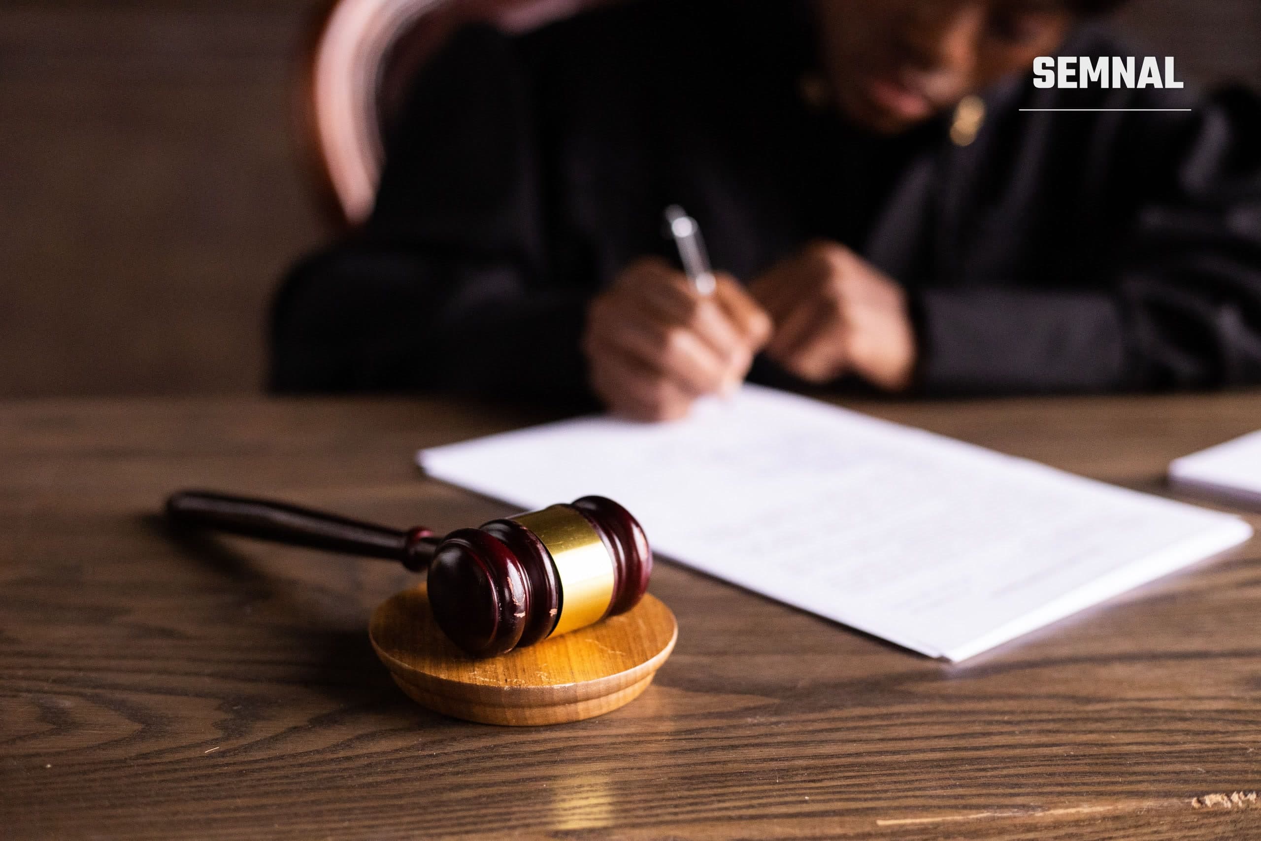 Gavel de judecător pe birou, în prim-plan, cu o persoană semnând documente în fundal, imagine ilustrativă pentru un dosar penal