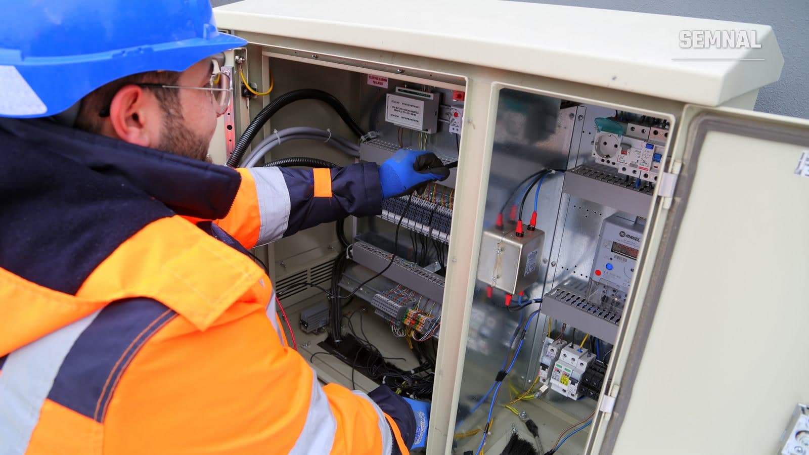 Electrician cu cască de protecție și vestă reflectorizantă verifică un tablou electric cu cabluri și relee, în timpul unei intervenții tehnice