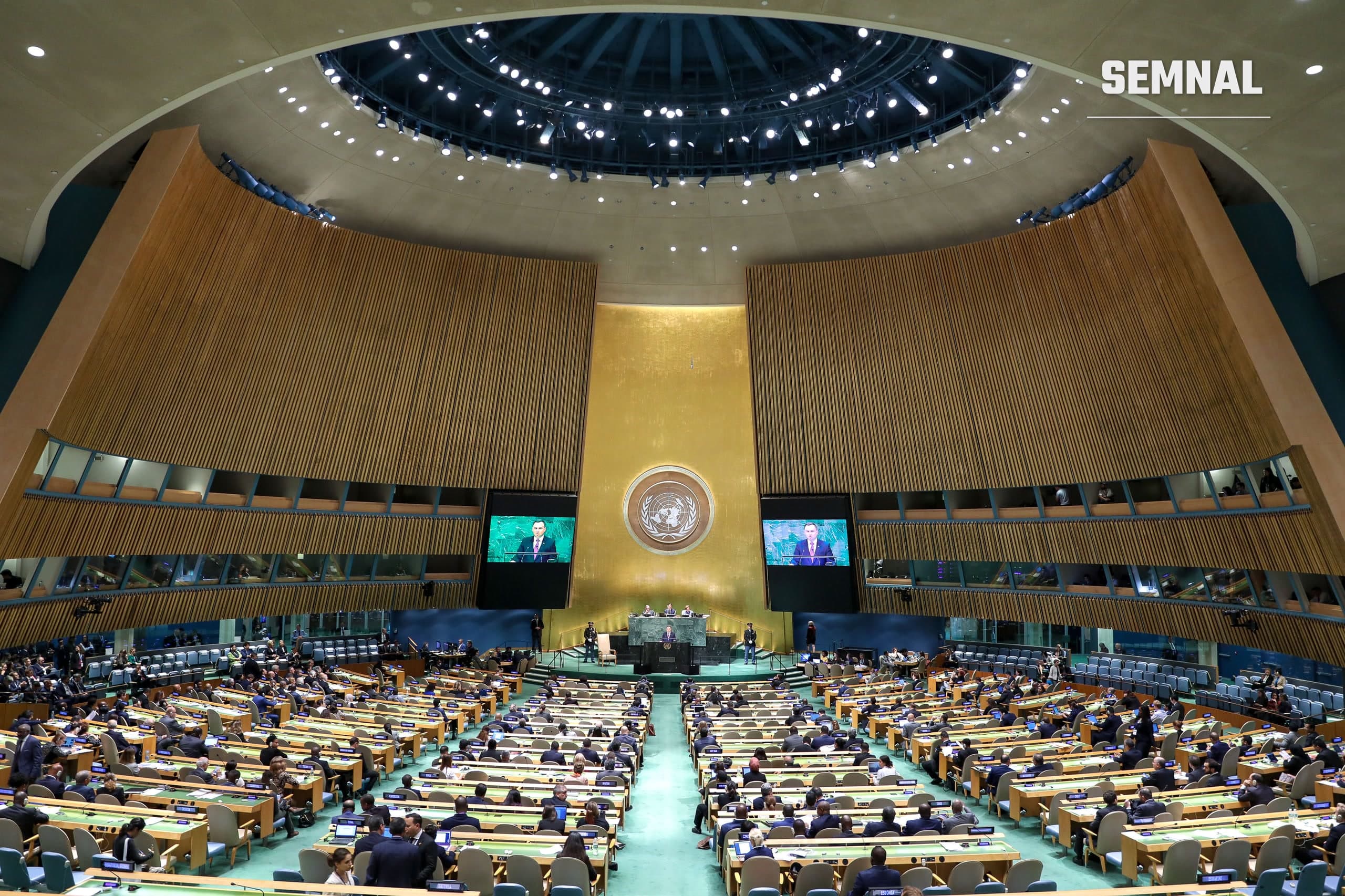 Sala Adunării Generale a ONU, la New York, plină cu delegați, în timpul unei sesiuni în care un vorbitor se adresează Adunării de la tribună, cu ecrane laterale active.