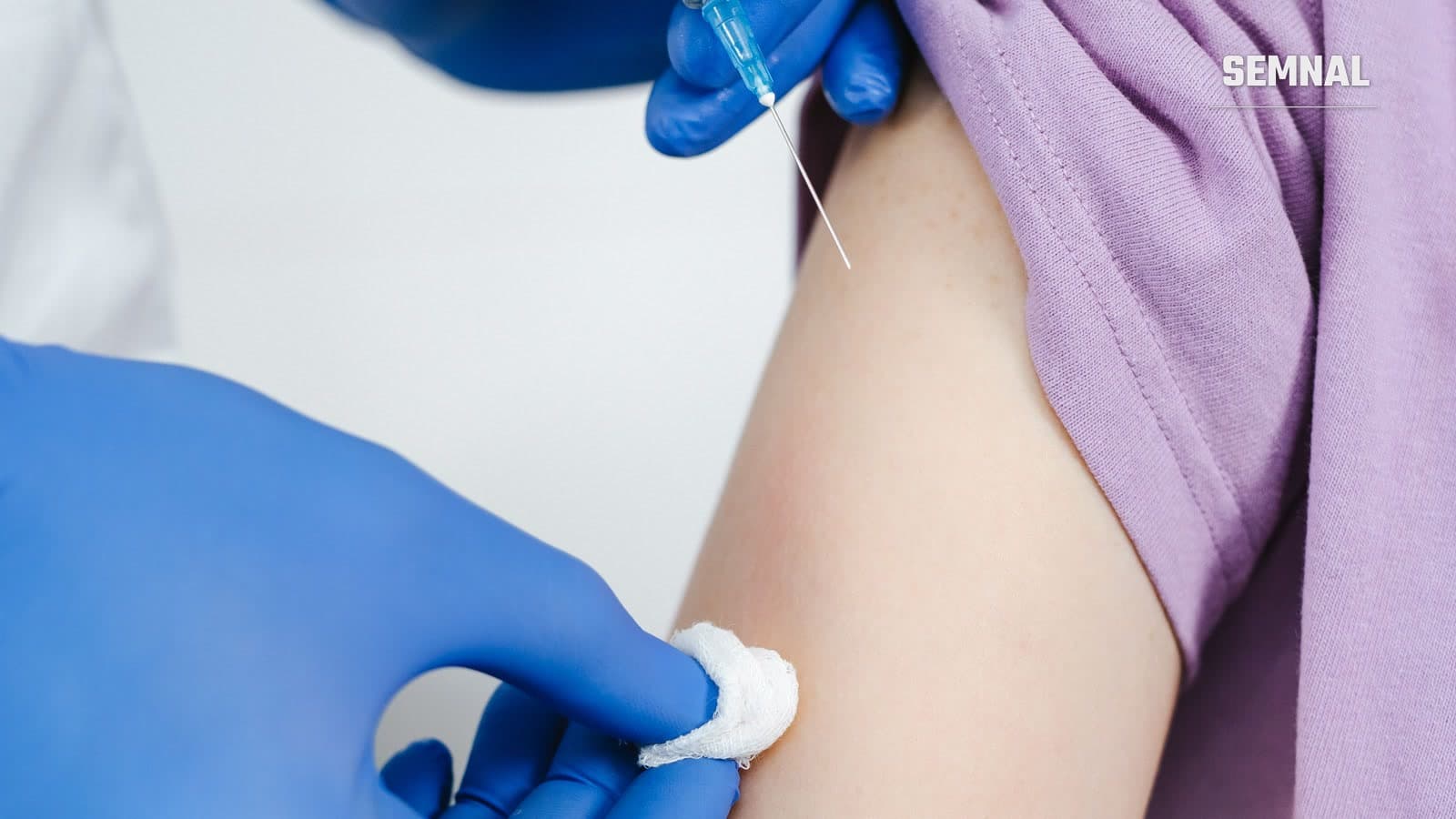 Cadru medical în mănuși albastre pregătește injecția cu seringa în brațul unui pacient, cu un tampon de tifon în cealaltă mână