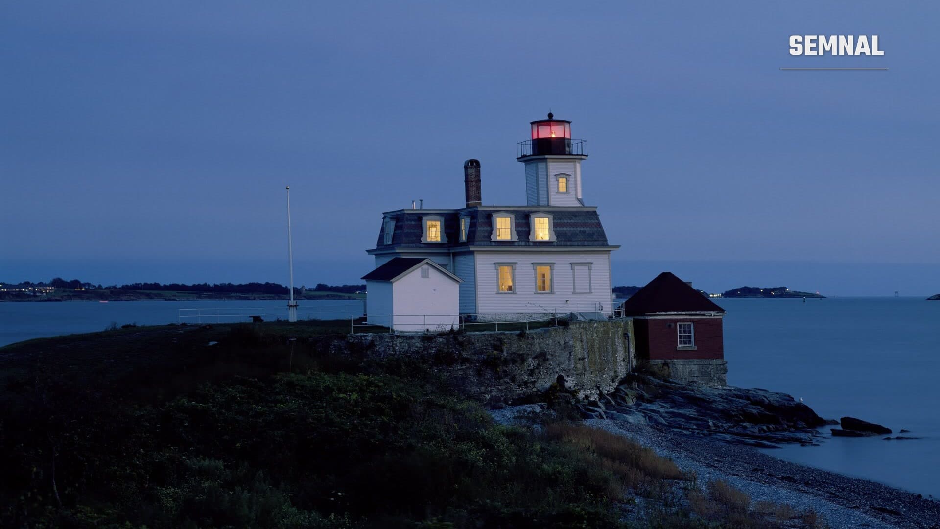 Farul Rose Island din Newport, Rhode Island, cu ferestrele luminate și felinarul roșu aprins, la amurg, pe o insulă din golful Narragansett