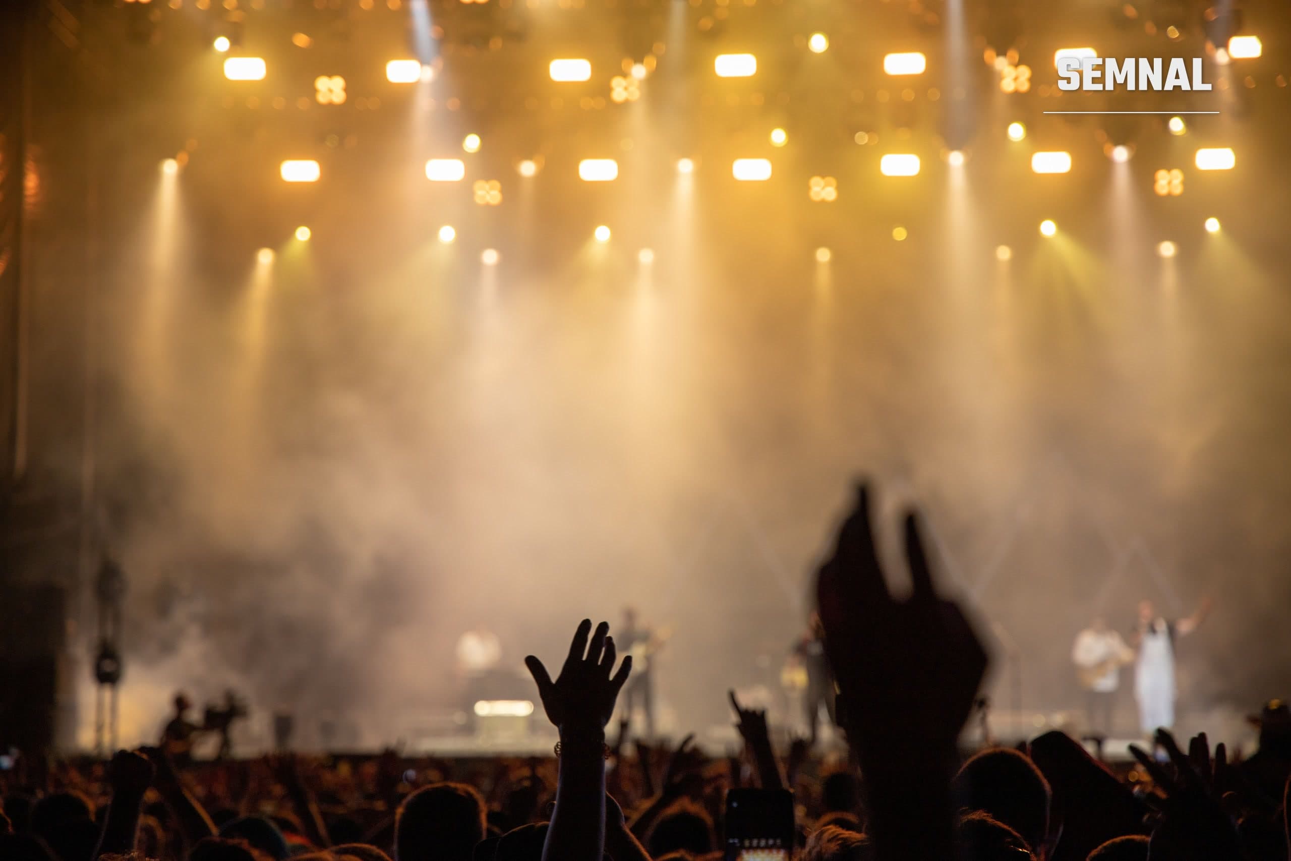 Public cu mâinile ridicate la un concert în aer liber, în fața unei scene luminate de reflectoare și învăluite în fum