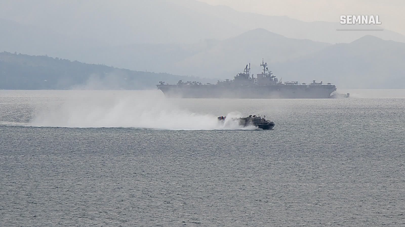 O navă de desant pe pernă de aer gonește pe apă, lăsând o dâră de spumă, cu nava de asalt amfibiu USS Makin Island în fundal, în fața unor munți învăluiți în ceață.