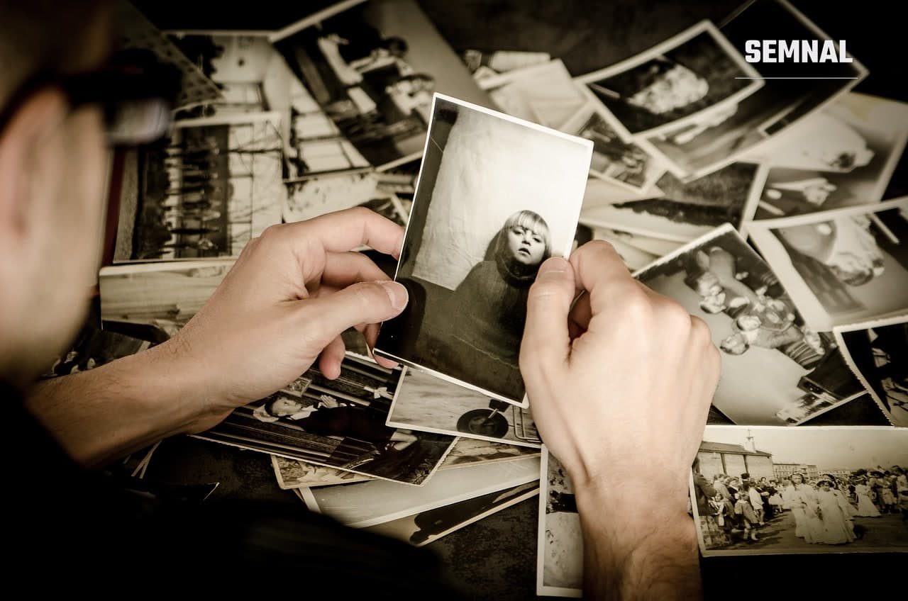 Un bărbat ține în mână o fotografie veche în alb-negru cu un copil, deasupra unui morman de alte poze vechi răsfirate pe masă. Foto jarmoluk / Pixabay