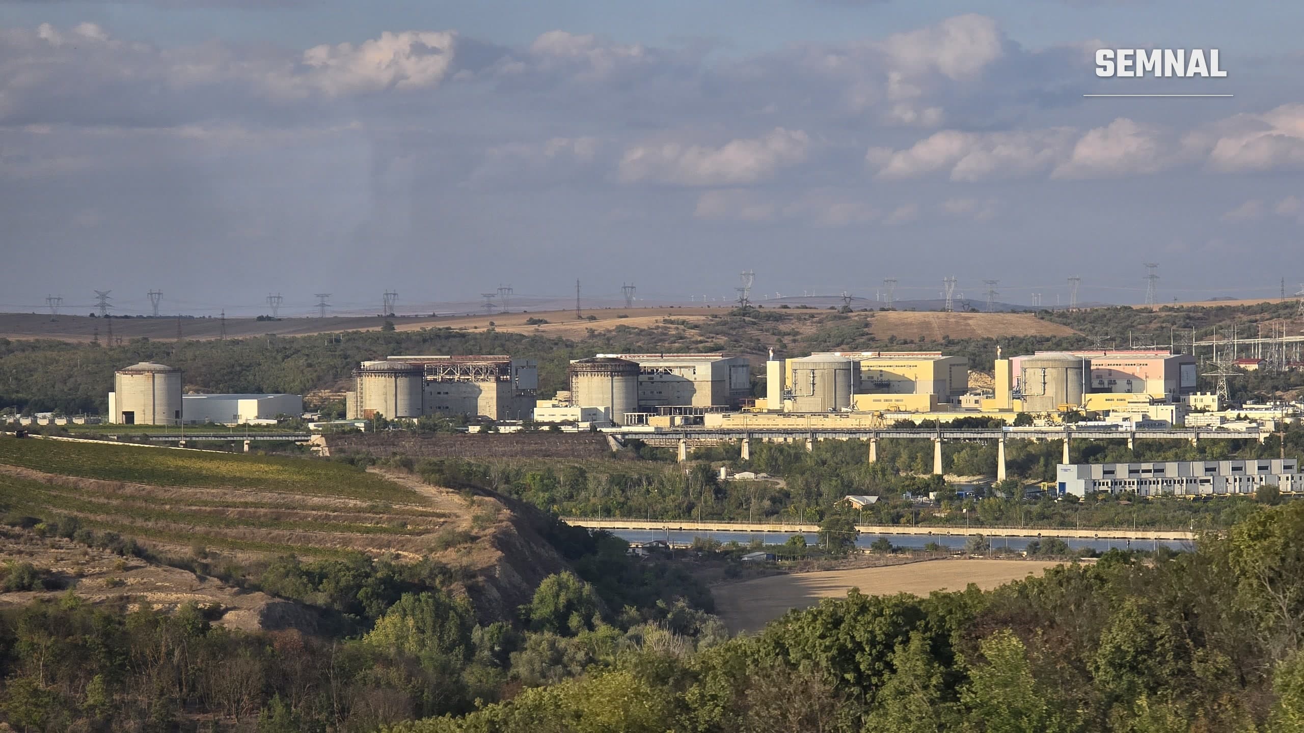 Vedere de ansamblu asupra centralei nucleare de la Cernavodă, cu clădirile reactoarelor și liniile electrice, văzută de pe dealurile din apropiere