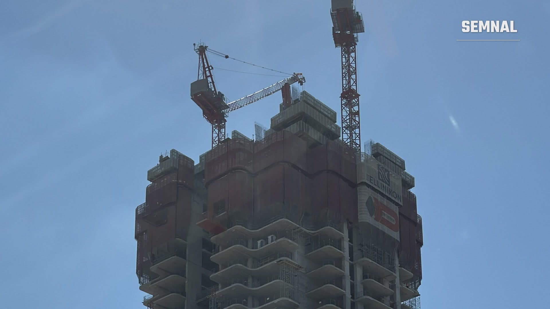 Partea superioară a turnului Riviera din Atena, în construcție, cu două macarale roșii și panoul Ellinikon pe fațadă, sub un cer senin