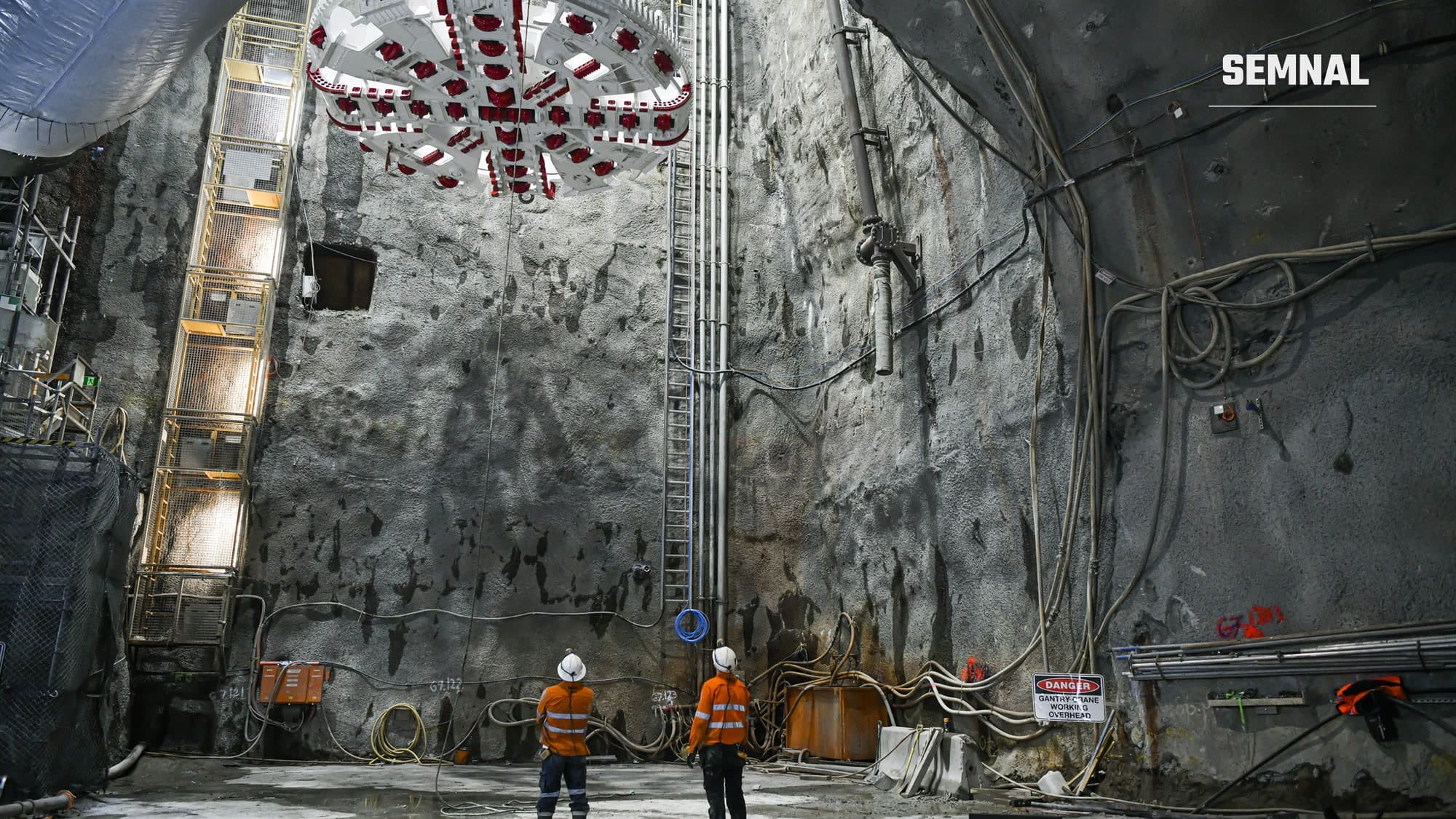 Capul de forare al unui utilaj TBM, suspendat deasupra unui puț de excavație, privit de doi muncitori în echipament de protecție