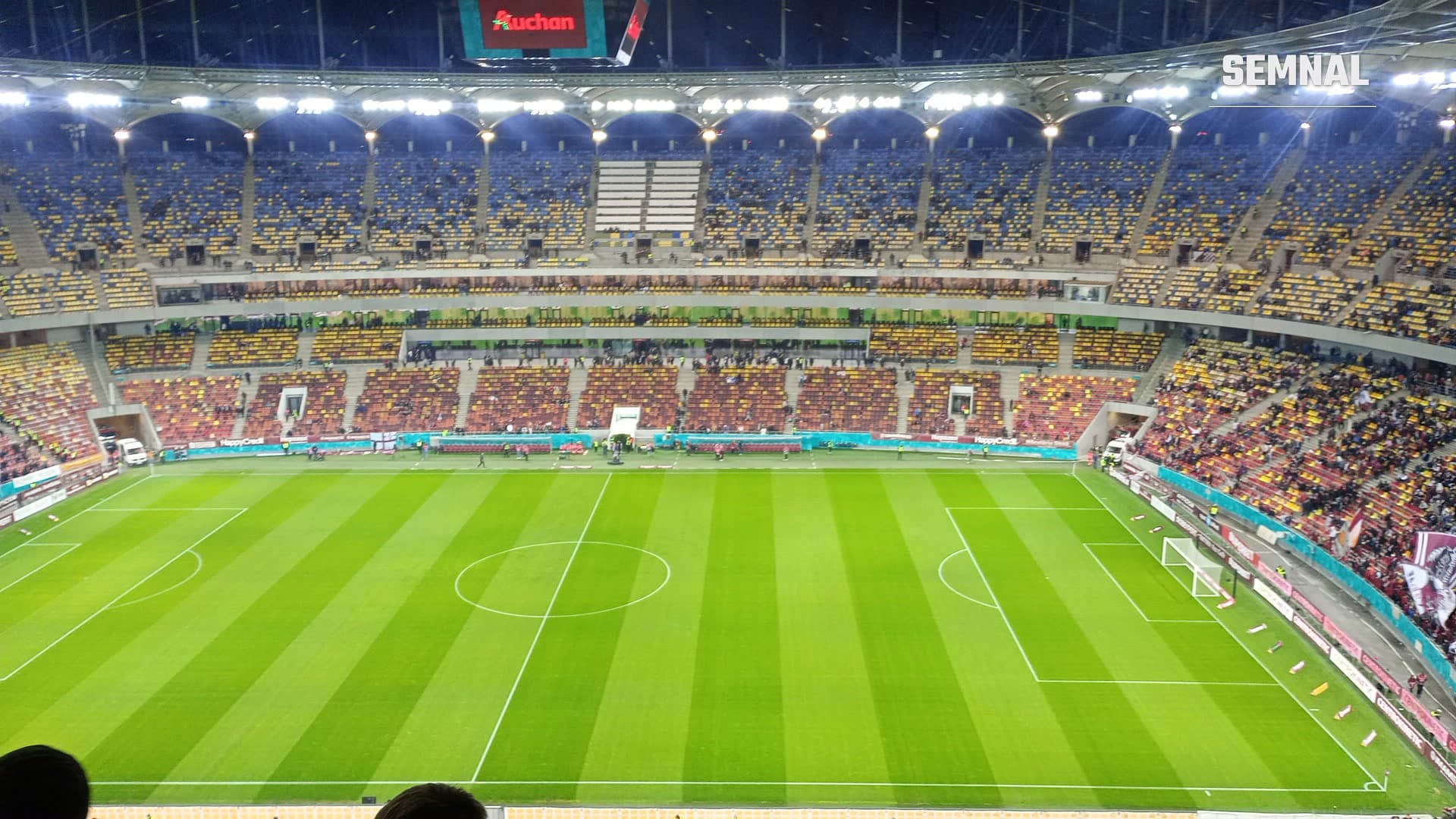 Arena Națională din București văzută din tribune înaintea unui meci de fotbal, cu gazon în dungi, tribune parțial ocupate și reflectoare aprinse