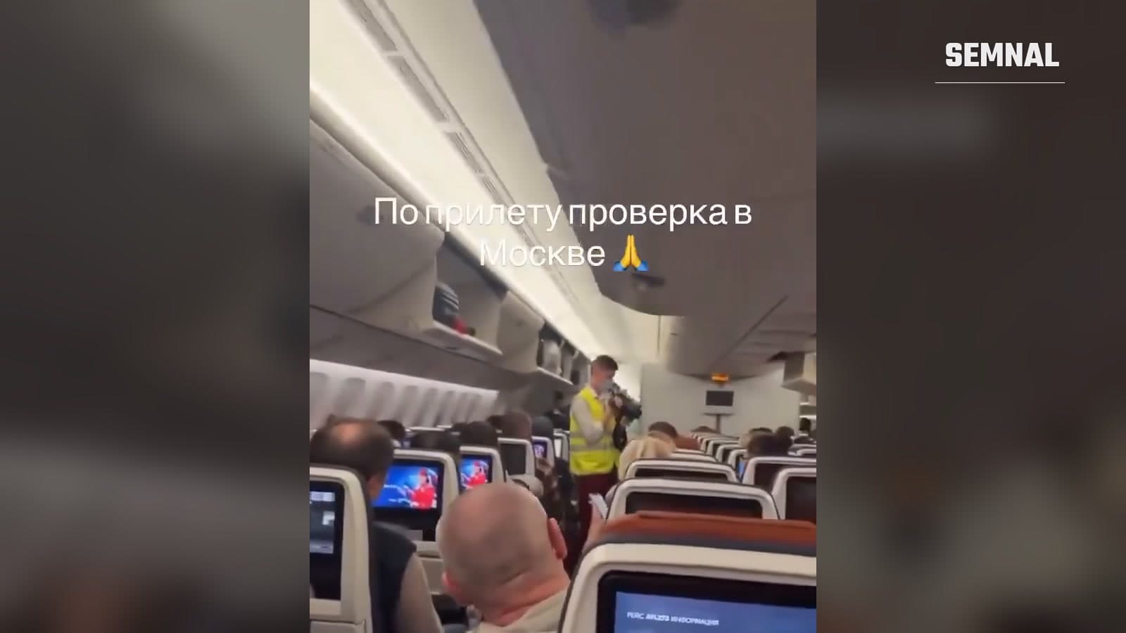 Pasageri așezați într-un avion de linie, în timp ce un bărbat cu vestă reflectorizantă și mască filmează pe culoarul dintre scaune