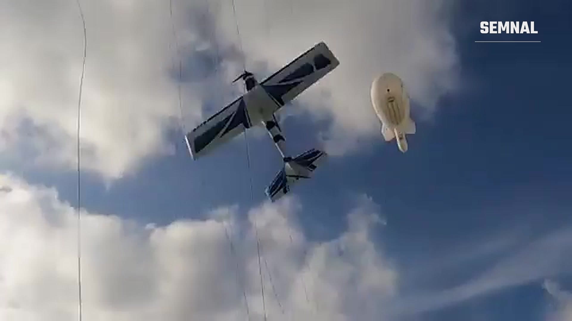 Dronă cu aripi fixe agățată în cablurile unui baraj de baloane, cu un aerostat alb pe cerul înnorat
