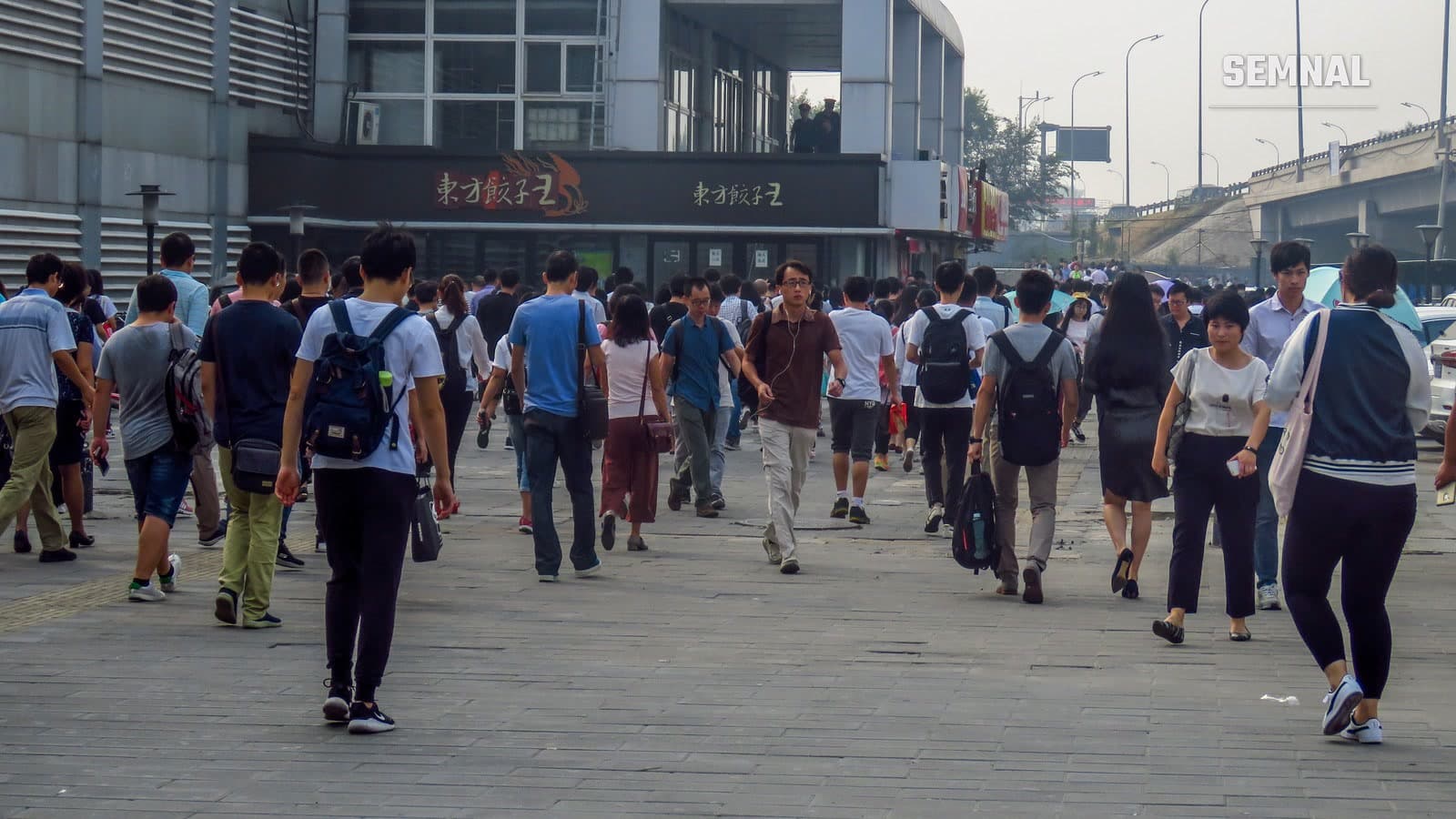 Mulțime de pietoni care merg pe o esplanadă pavată, în mare parte din spate sau din profil, în fața stației Xi'erqi din Beijing.