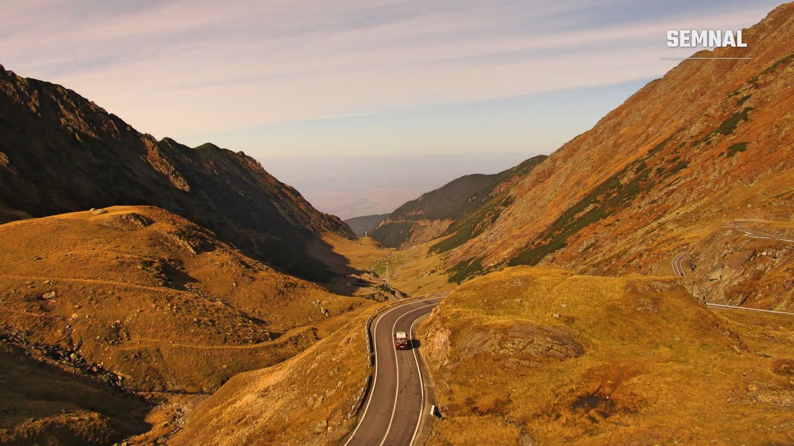 Drumul Transfăgărășan serpuiește printr-o vale din Munții Făgăraș, cu pante în nuanțe de ocru și auriu, o mașină pe asfalt și cer senin la orizont