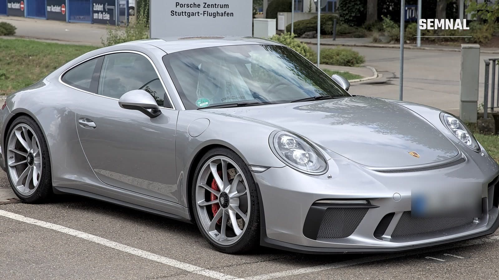 Porsche 911 GT3 argintiu, cu etriere roșii, văzut din față și din lateral, parcat lângă o indicație Porsche Zentrum Stuttgart