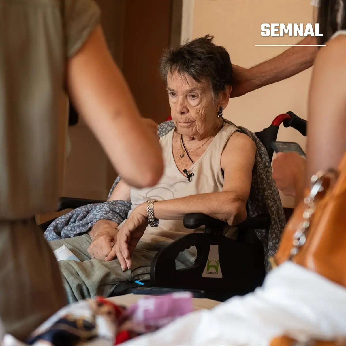 Femeie vârstnică cu părul scurt și închis la culoare, așezată într-un scaun cu rotile, cu un microfon mic prins de bluză, înconjurată de oameni într-o încăpere
