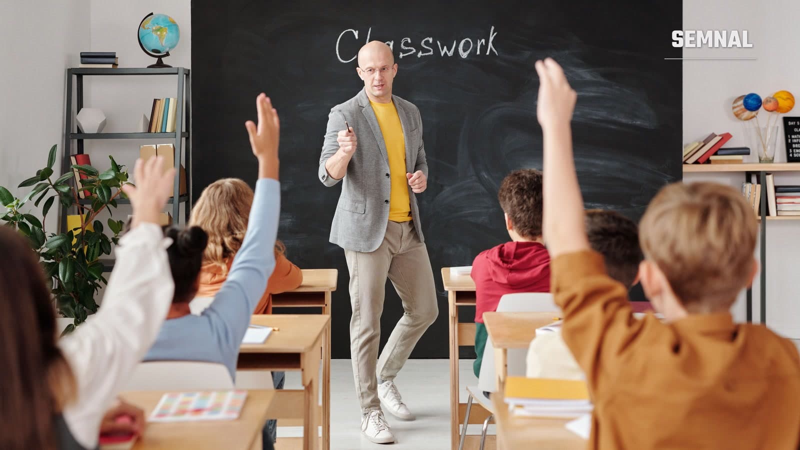 Profesor în fața unei table negre, într-o sală de clasă, în timp ce mai mulți elevi, văzuți din spate, ridică mâna să răspundă