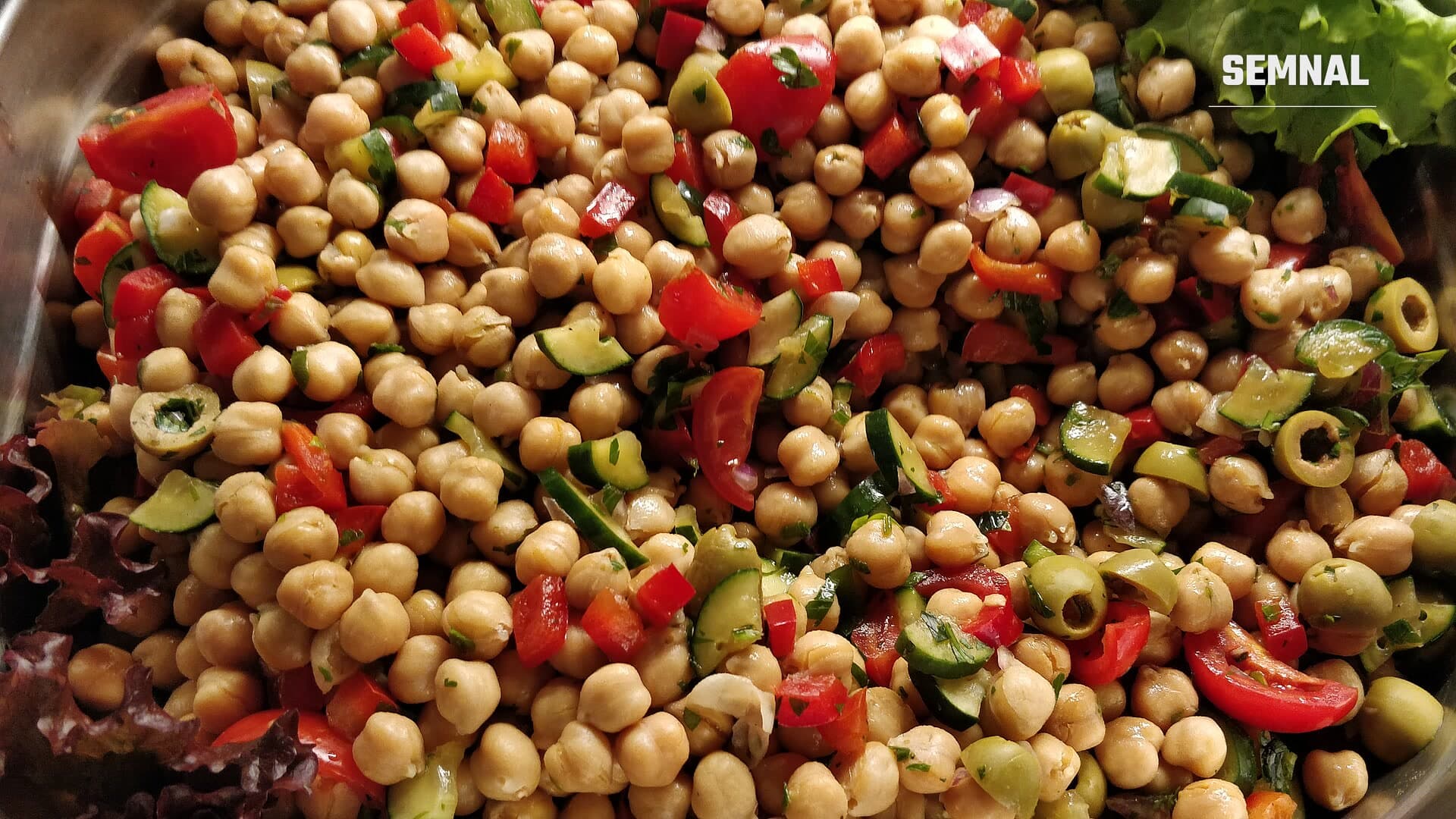 Salată de năut cu cuburi de roșii, ardei roșu, felii de castravete și măsline verzi, cu frunze de salată verde la margine, într-un vas de sticlă
