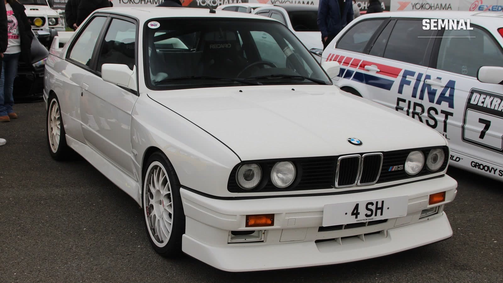 BMW M3 E30 alb, prima generație a modelului, fotografiat din față-lateral pe asfalt la un eveniment auto