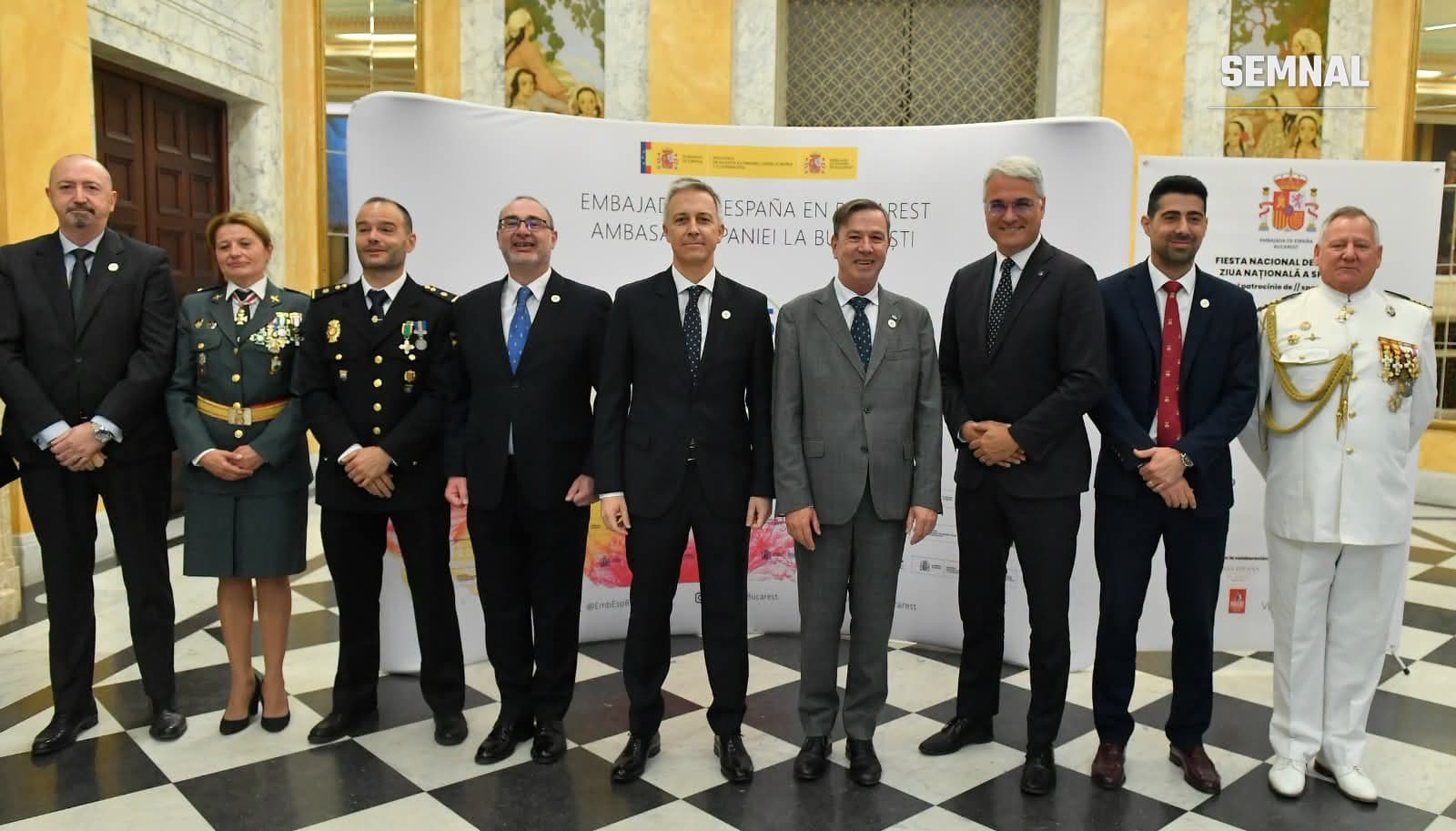 Nouă persoane, printre care ambasadorul Spaniei în România, în costume și uniforme de gală, pozează în fața panoului Ambasadei Spaniei la București, într-un hol cu pardoseală în carouri alb-negru.