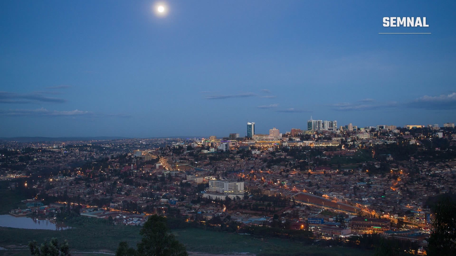 Panoramă a orașului Kigali, capitala Rwandei, la amurg, cu clădirile din centru luminate și luna pe cerul albastru