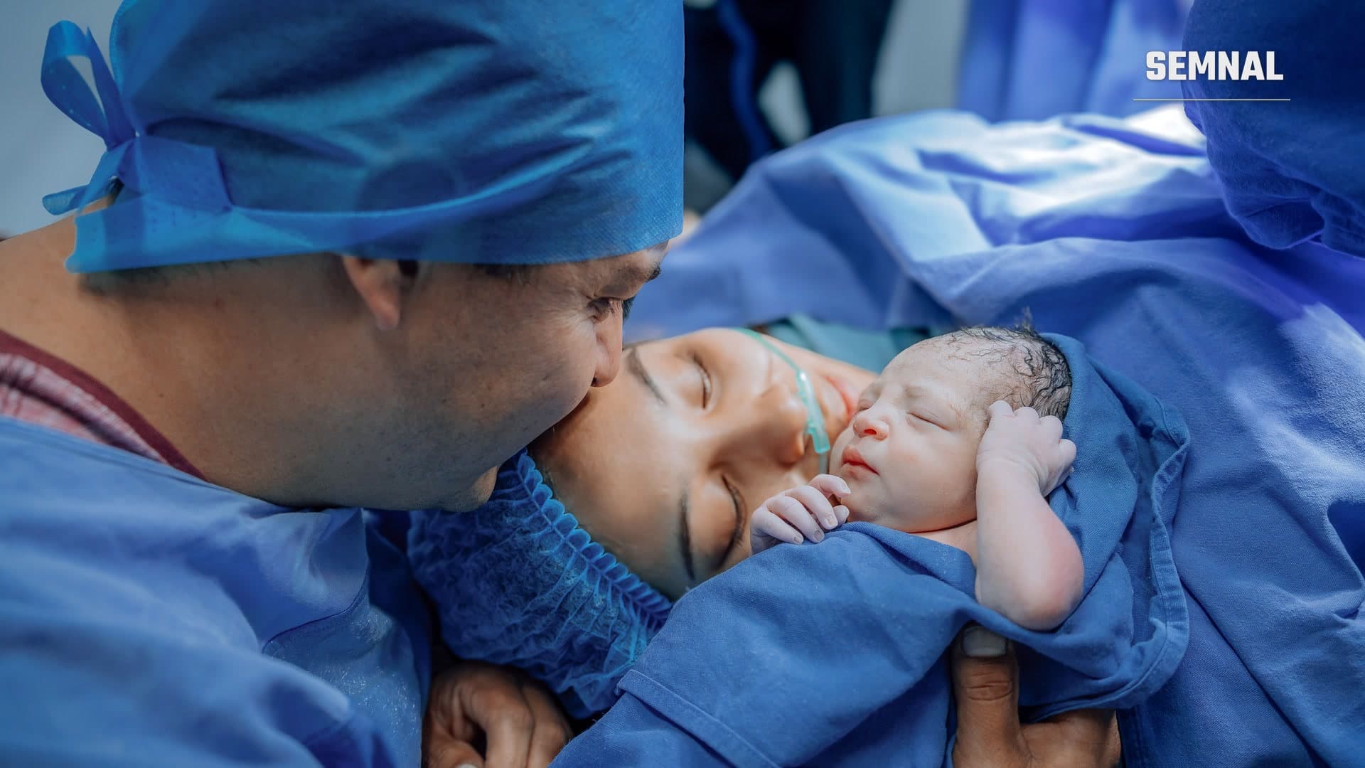 Un bărbat în echipament chirurgical își sărută partenera care ține pe piept un nou-născut, în sala de nașteri
