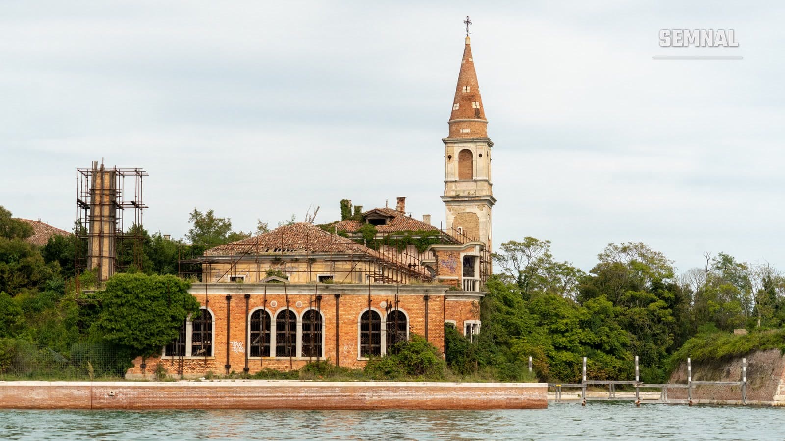 Clopotnița de cărămidă și clădirea în ruină a fostului spital de pe insula Poveglia, văzute din laguna Veneției, între vegetație