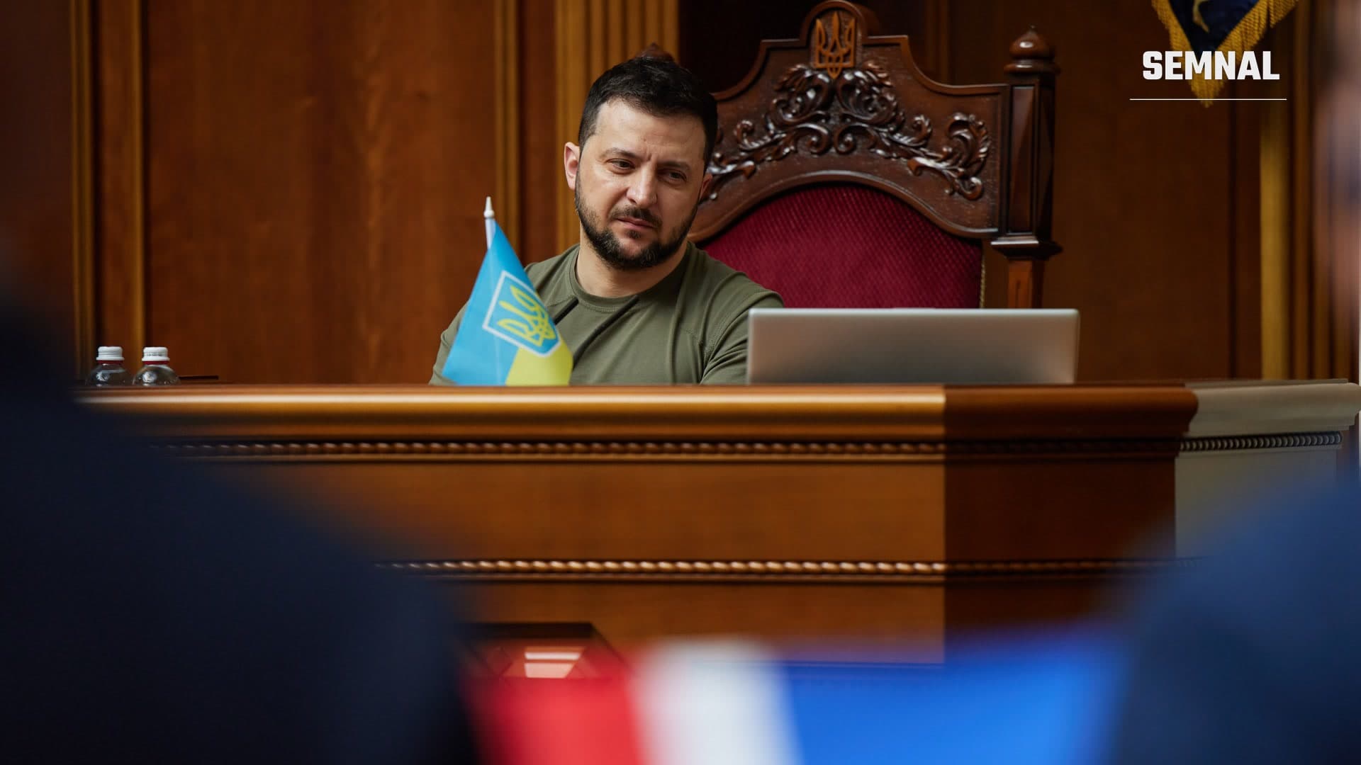 Volodimir Zelenski, în tricou kaki, așezat la un pupitru din lemn într-o ședință a Radei Supreme, cu un drapel mic al Ucrainei în față. (Foto: Oficiul Președintelui Ucrainei, CC0)
