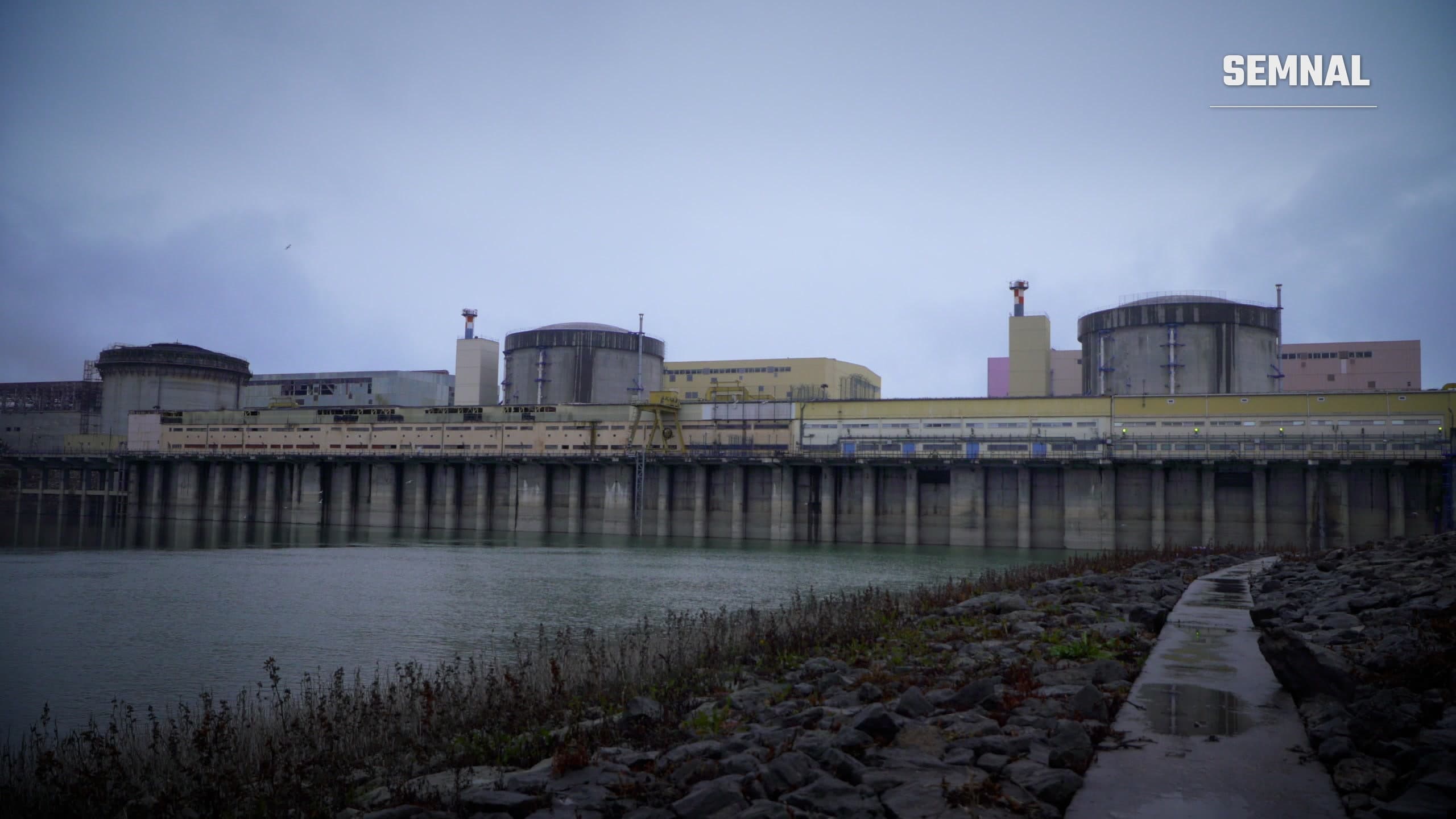 Centrala nucleară de la Cernavodă văzută de pe malul apei, cu clădirile reactoarelor și digul de piatră în prim-plan, într-o zi înnorată. Foto: IAEA Imagebank, CC BY 2.0