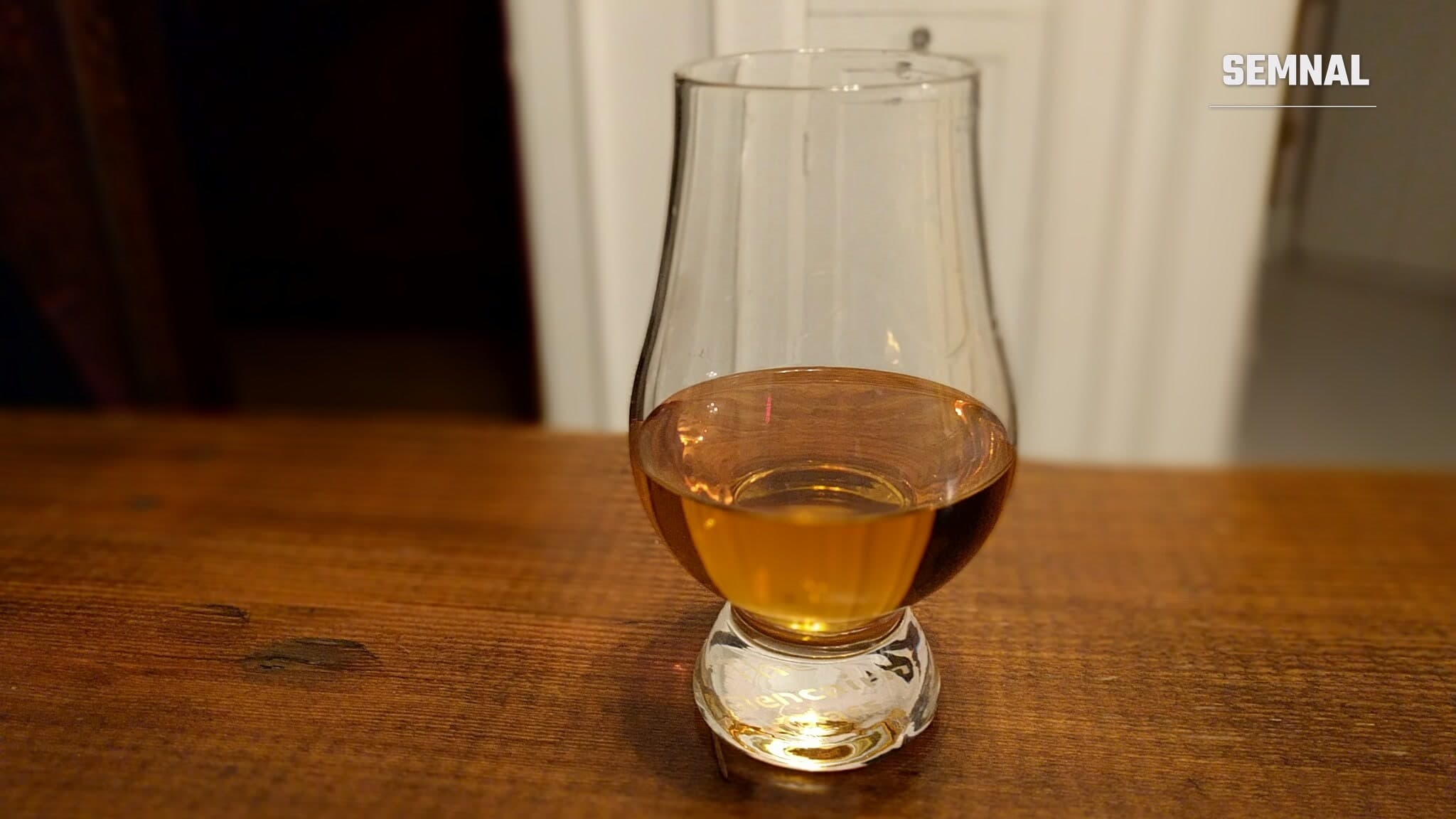 Pahar Glencairn umplut pe jumătate cu whisky chihlimbariu, așezat pe o masă de lemn, într-o încăpere slab luminată