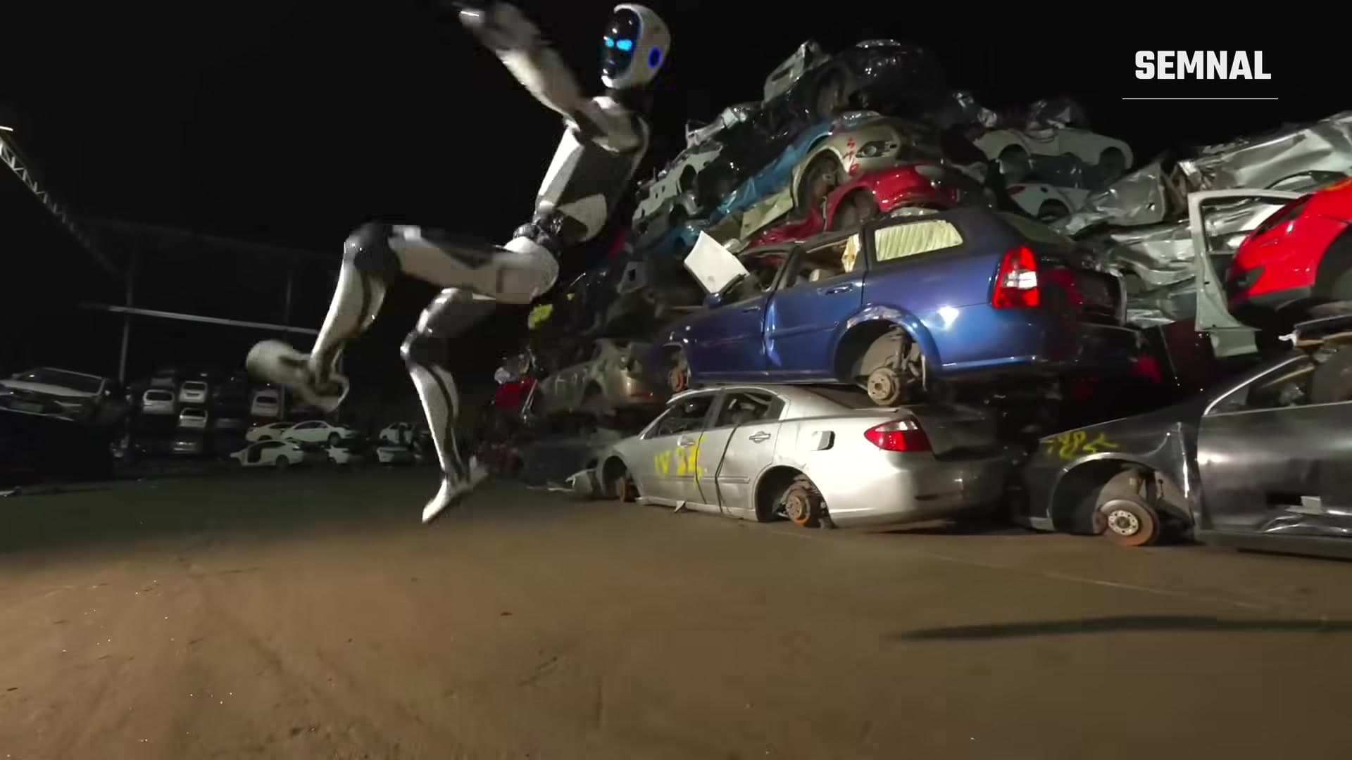 Robotul umanoid Unitree H2-X execută o lovitură cu piciorul lângă un morman de autoturisme casate, noaptea