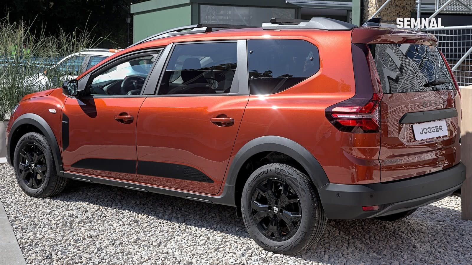 Dacia Jogger portocaliu, văzut din lateral-spate, cu jante negre și plăcuța cu numele modelului pe hayon, expus pe un platou cu pietriș la IAA 2021