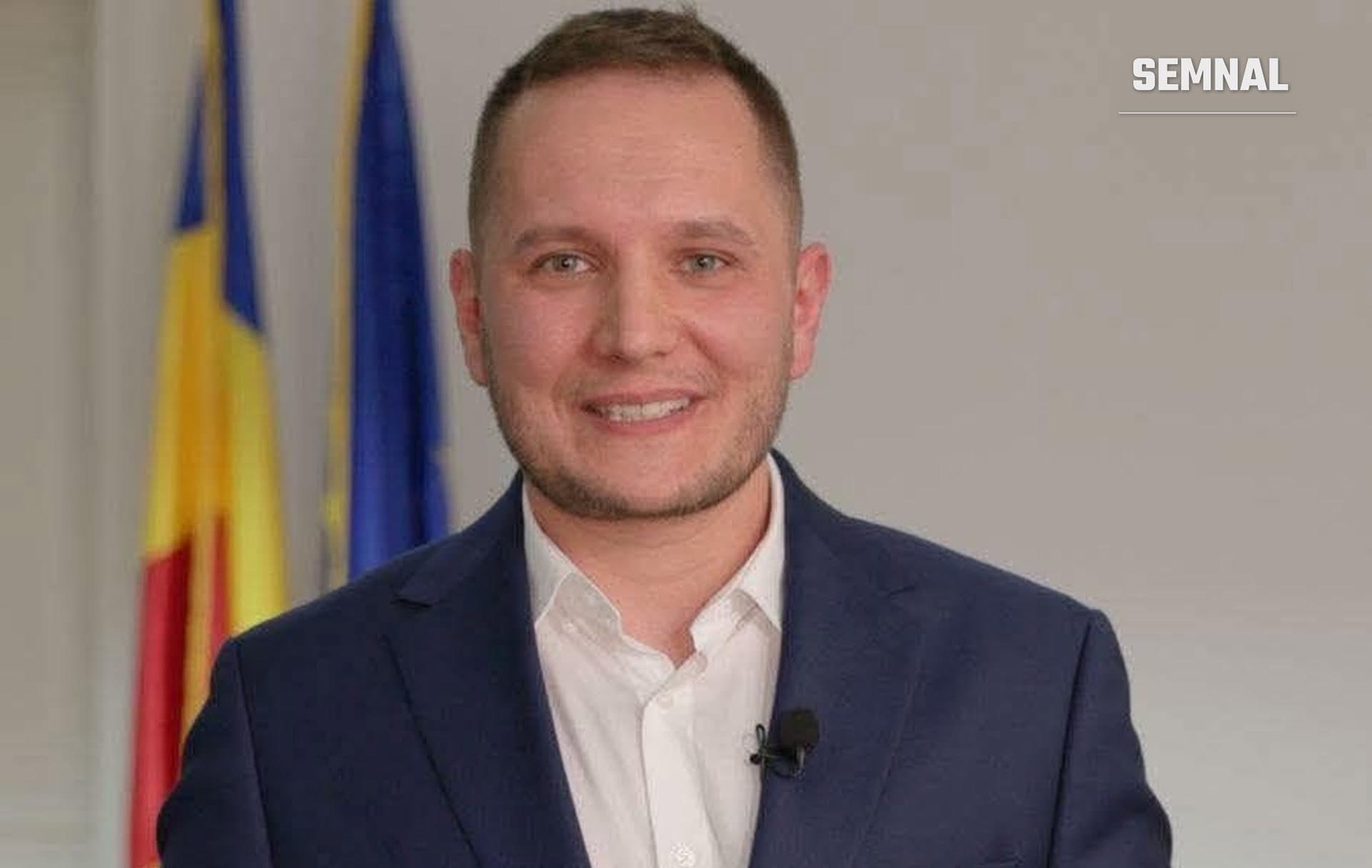 Andrei Nistor, zâmbind, în costum bleumarin și cămașă albă, cu microfon la rever, în fața drapelelor României și Uniunii Europene